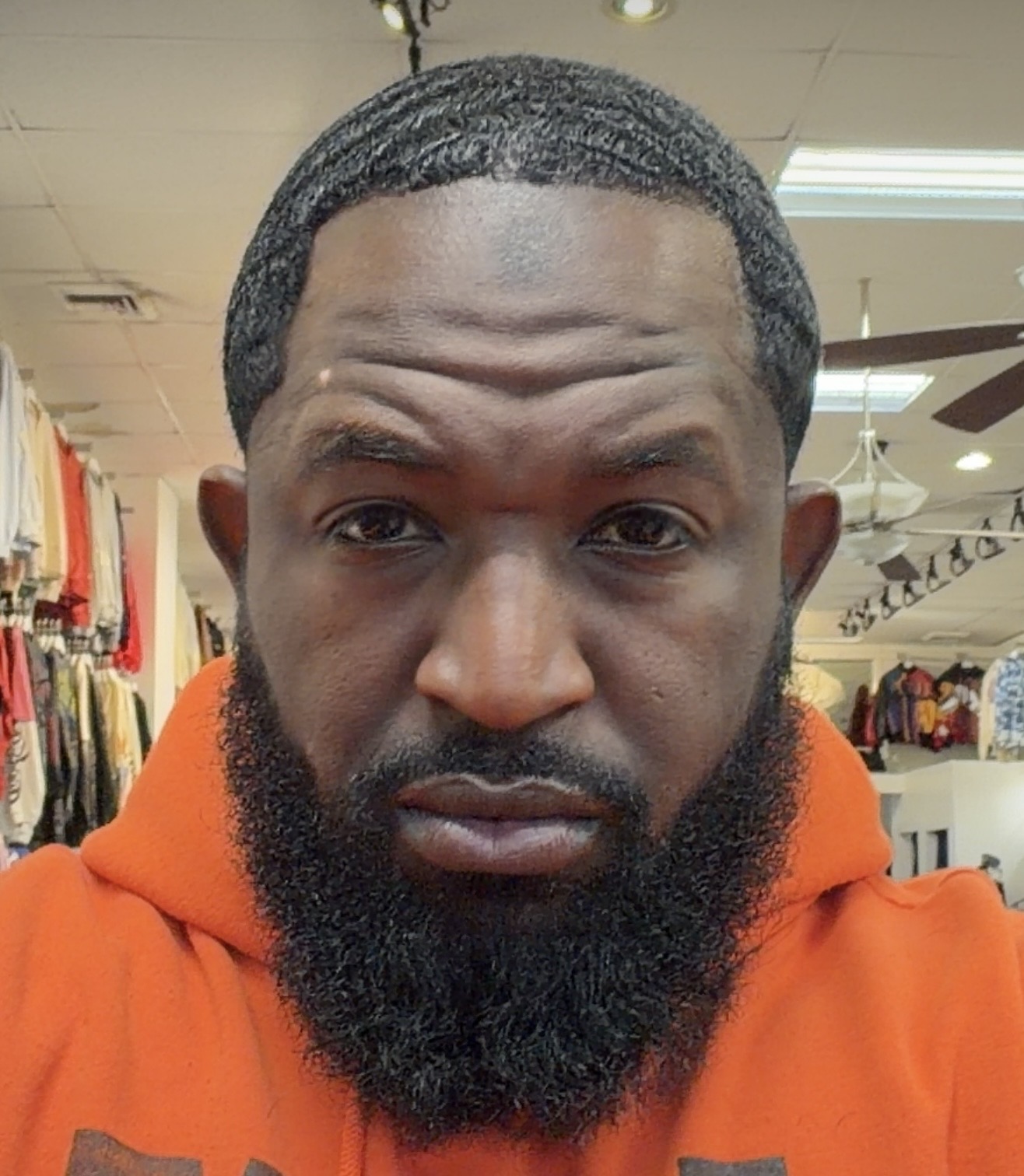 Man with a beard wearing an orange hoodie in a store, looking directly at the camera.