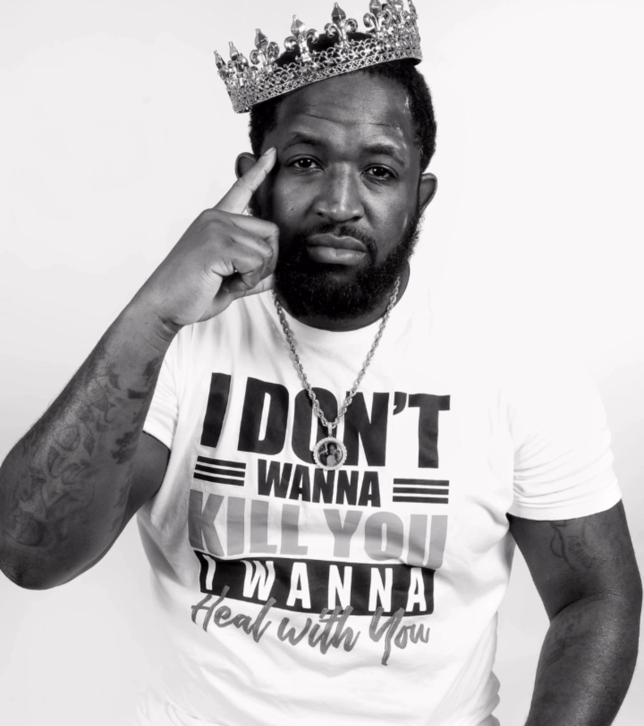 Man wearing a crown, pointing to his temple, with a T-shirt that has text, against a plain background.