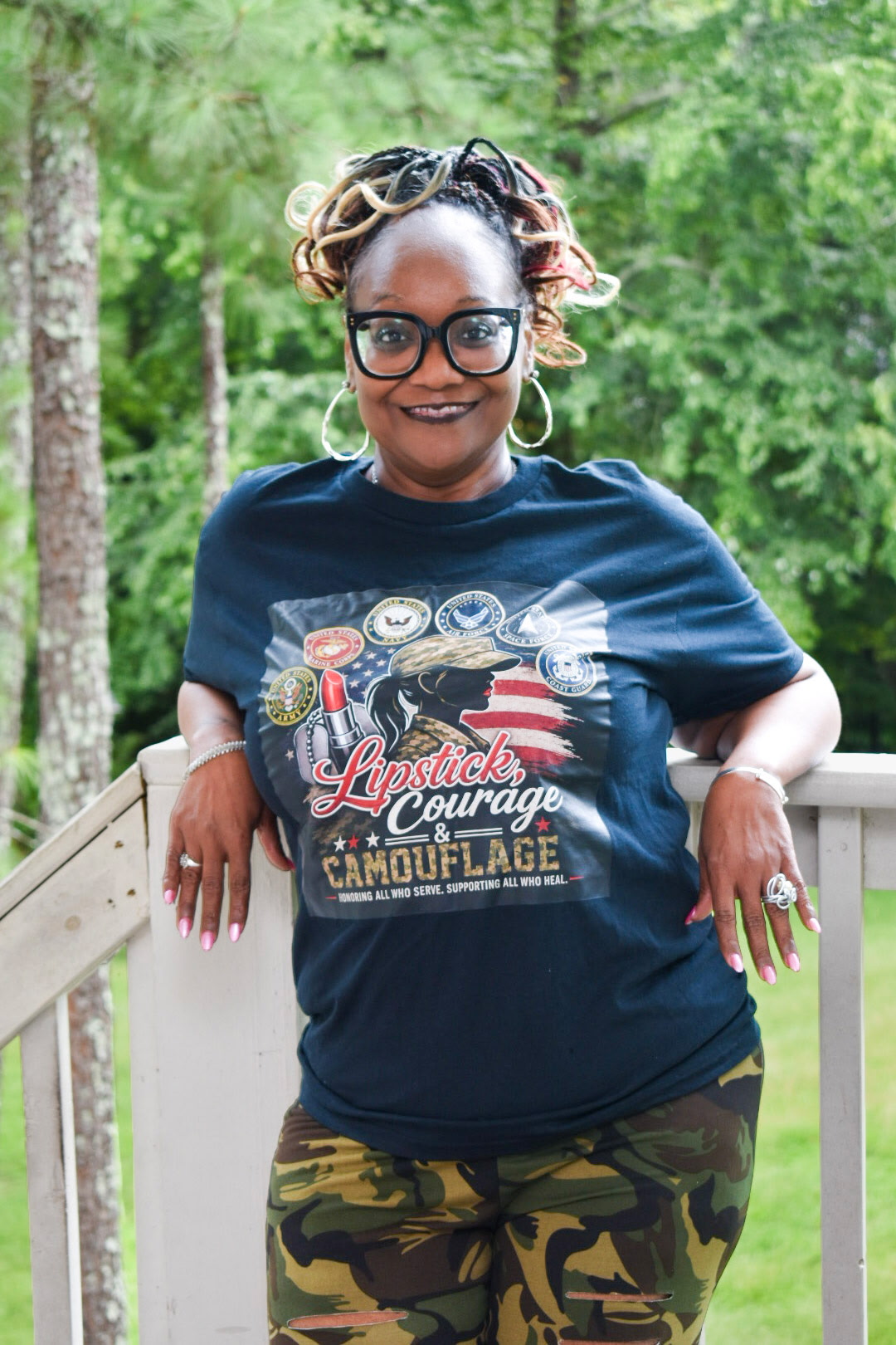 Woman with glasses and styled hair smiling outdoors, leaning on a wooden railing, wearing a graphic T-shirt and camouflage pants.
