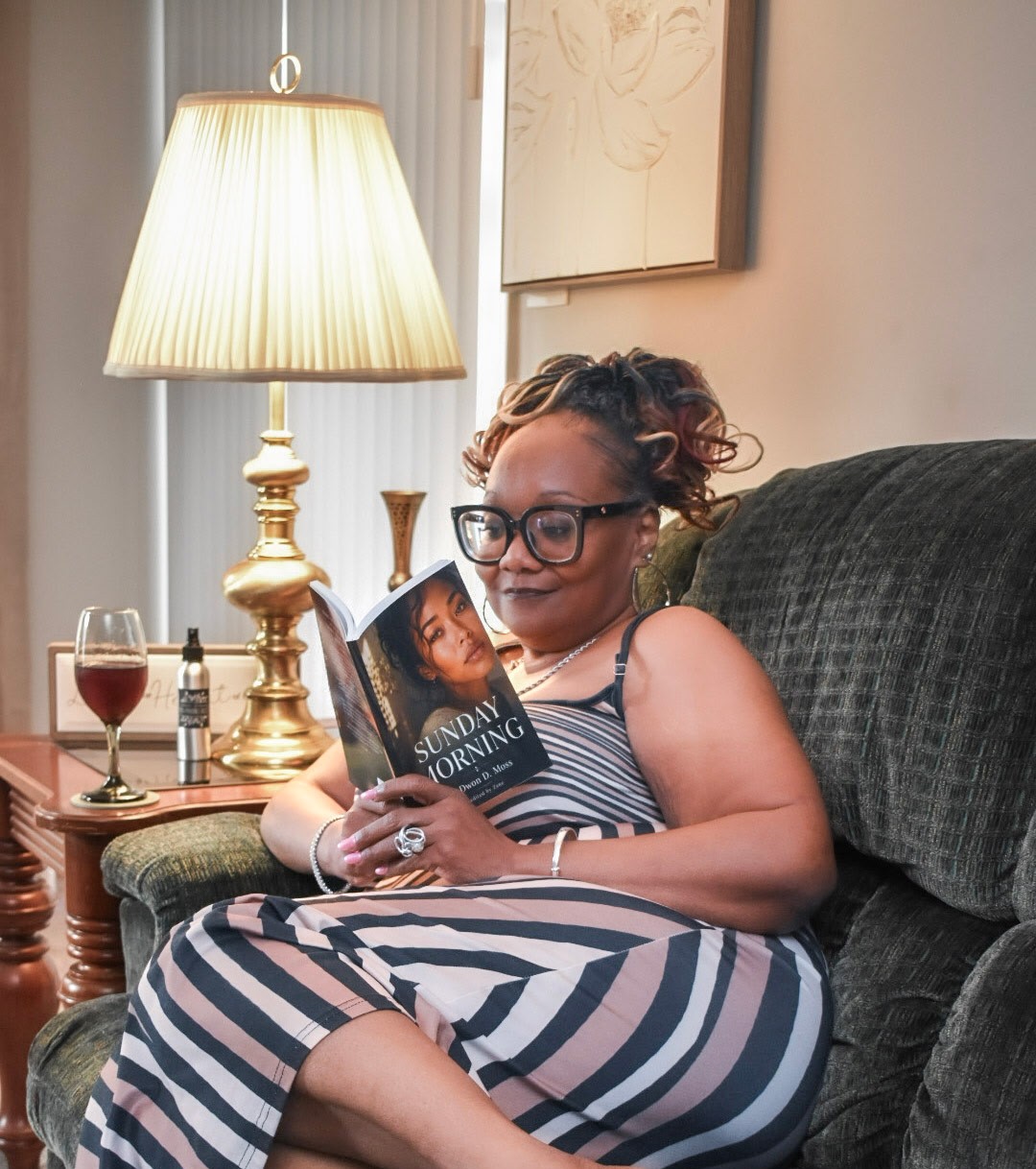 Woman with glasses sitting on a sofa, reading a magazine, with a lamp and wine glass nearby.