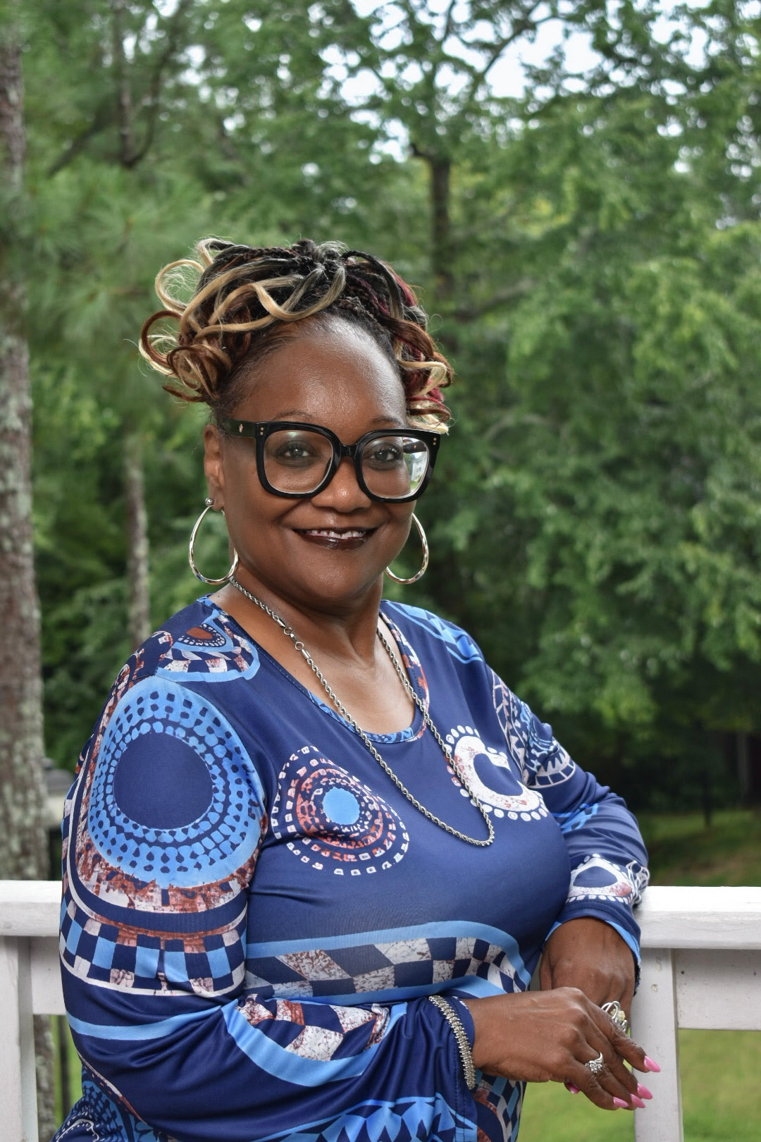 Woman with glasses and earrings outdoors, smiling, wearing a blue patterned dress, with trees in the background.