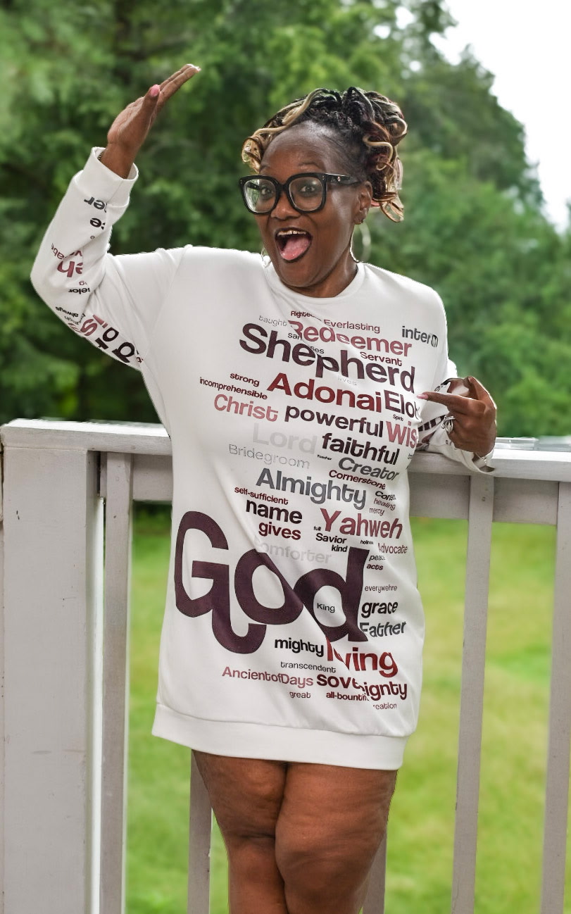 Woman outdoors on a porch, smiling, wearing glasses and a white oversized shirt with text, raising her right hand.