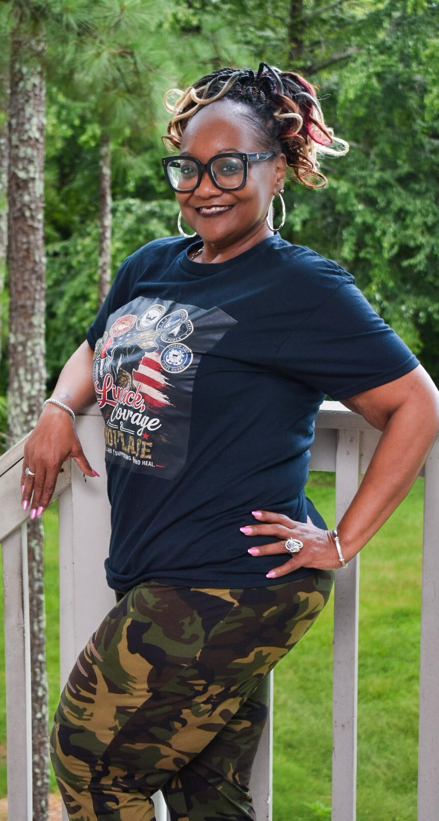 Woman with glasses and curly hair outdoors, smiling, wearing a navy T-shirt and camouflage pants, standing near a railing.