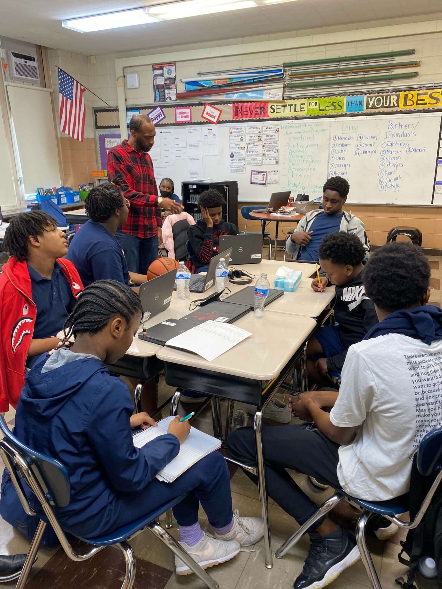 Students sit at desks in a classroom, engaging with a teacher and each other, with educational posters on the wall.