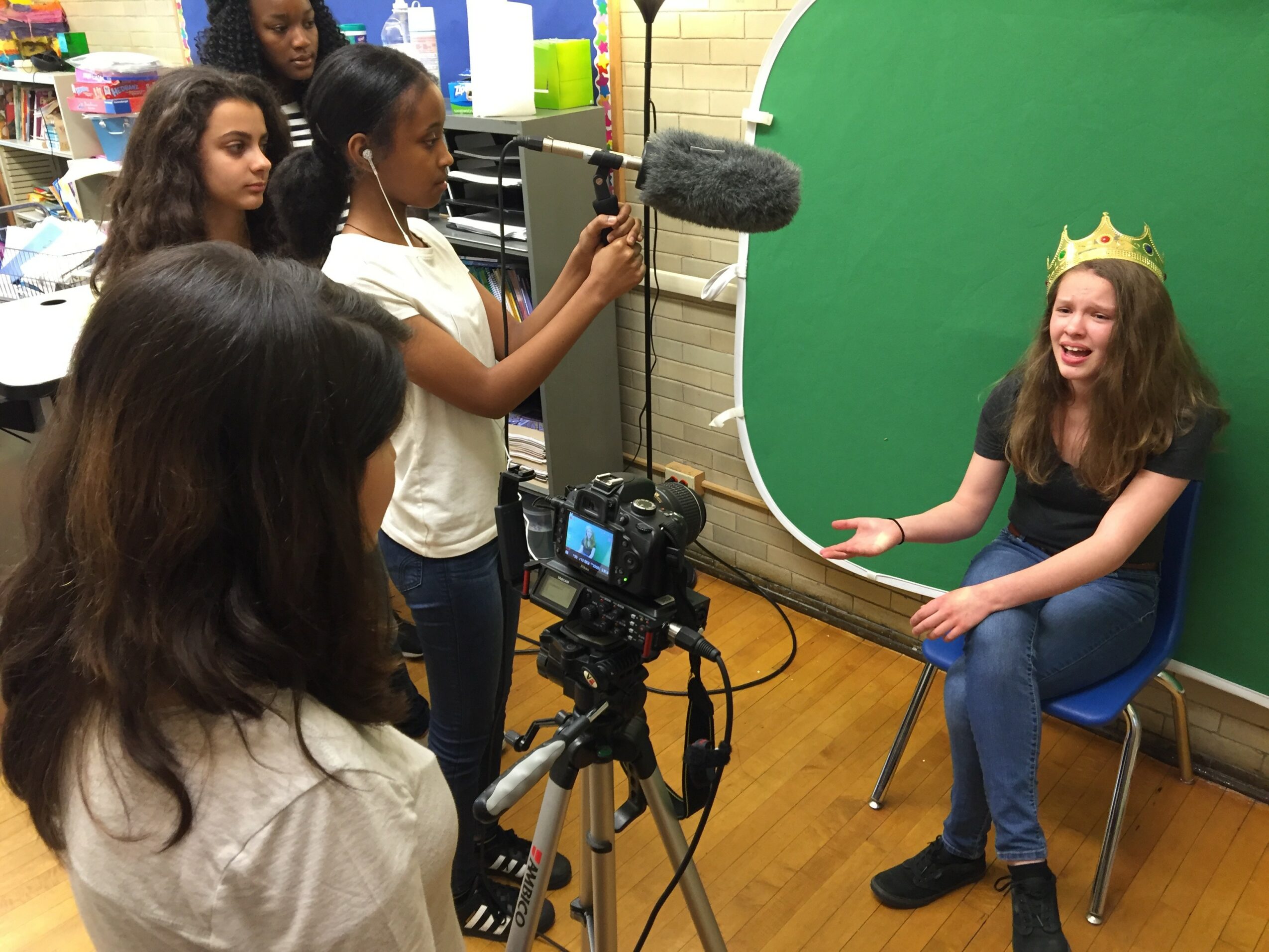 Group of children filming a girl wearing a crown sitting in front of a green screen in a classroom.