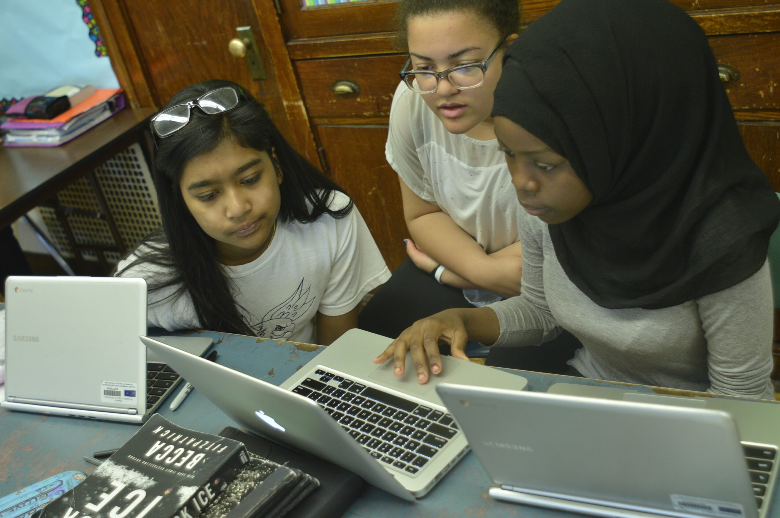 Three women sit at a table with laptops, engaged in a discussion or work, in a room with wooden furniture.