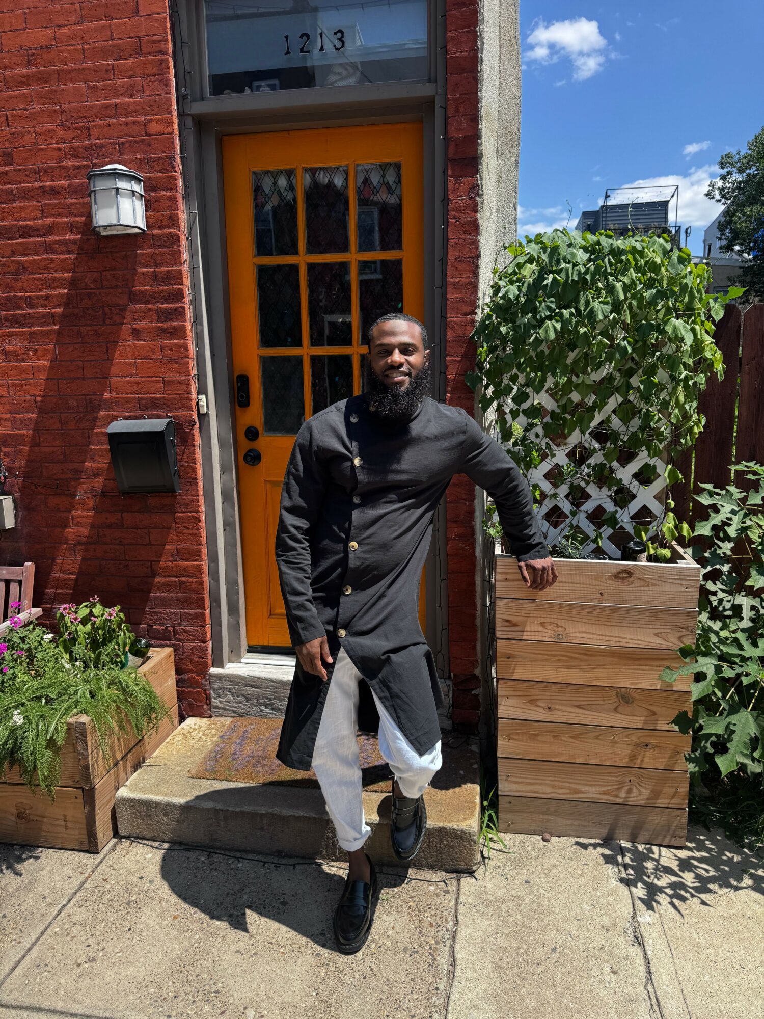 Person standing outside near a brick building with a yellow door, plants, and wooden planters, sunny day with blue sky.
