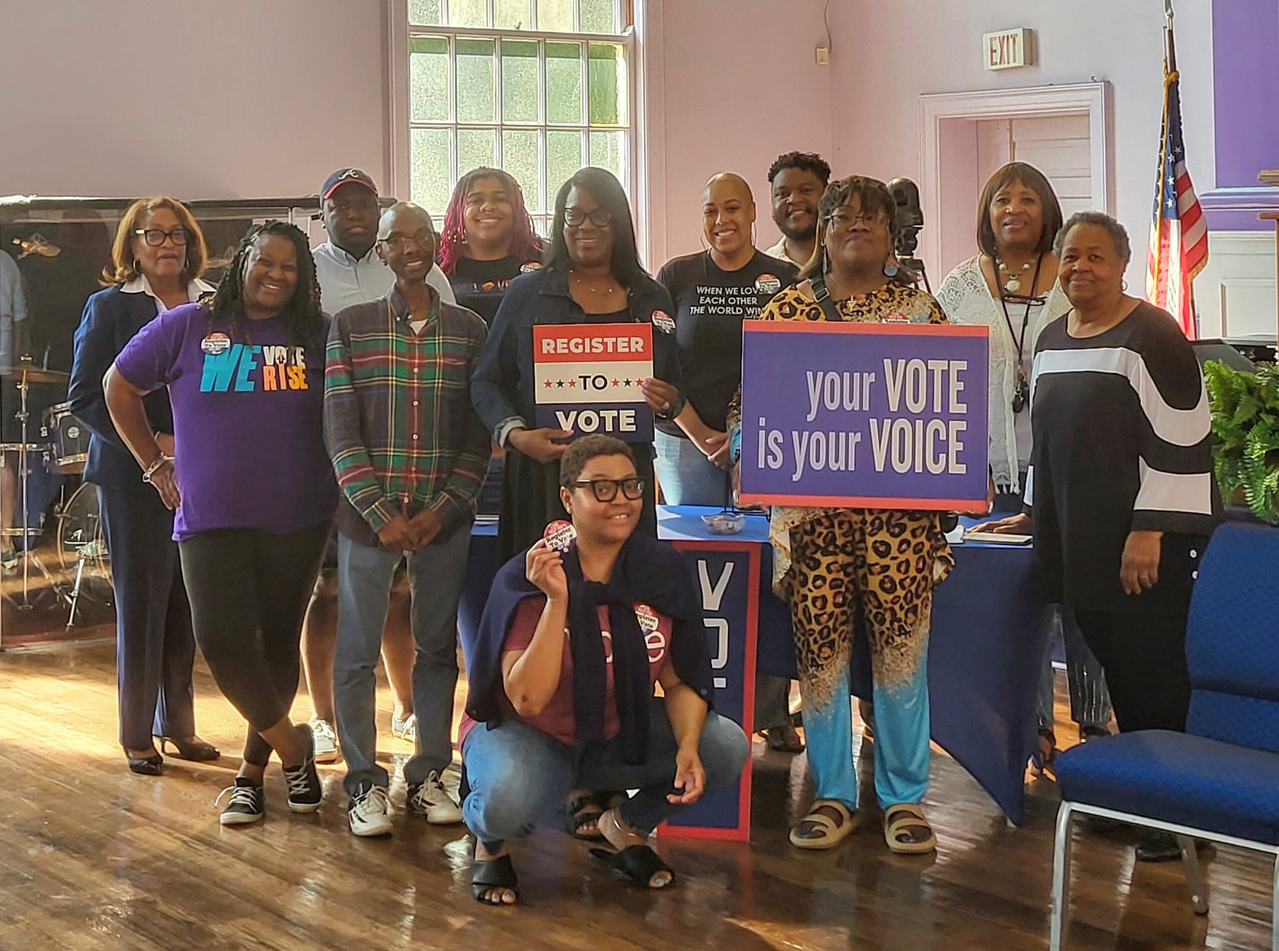 Group of people holding signs about voting, standing indoors with a window and American flag in background.