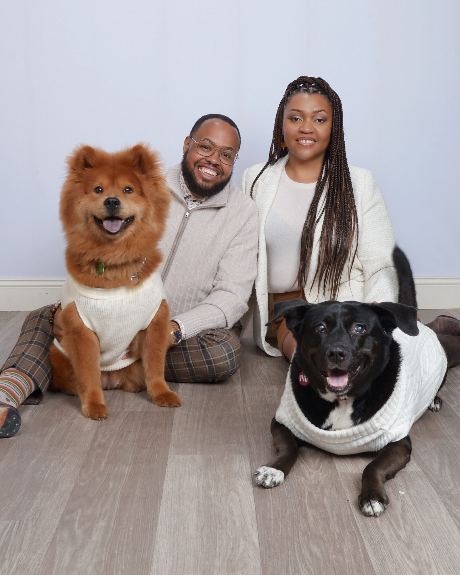 Two people and two dogs sitting on the floor against a light blue background, smiling. The person on the left is a man with glasses, and the person on the right is a woman with long braided hair. The dog on the left is a fluffy, brown dog, and the dog on the right is a black and white dog. Alt_text_word_count: 36, confidence: 0.95