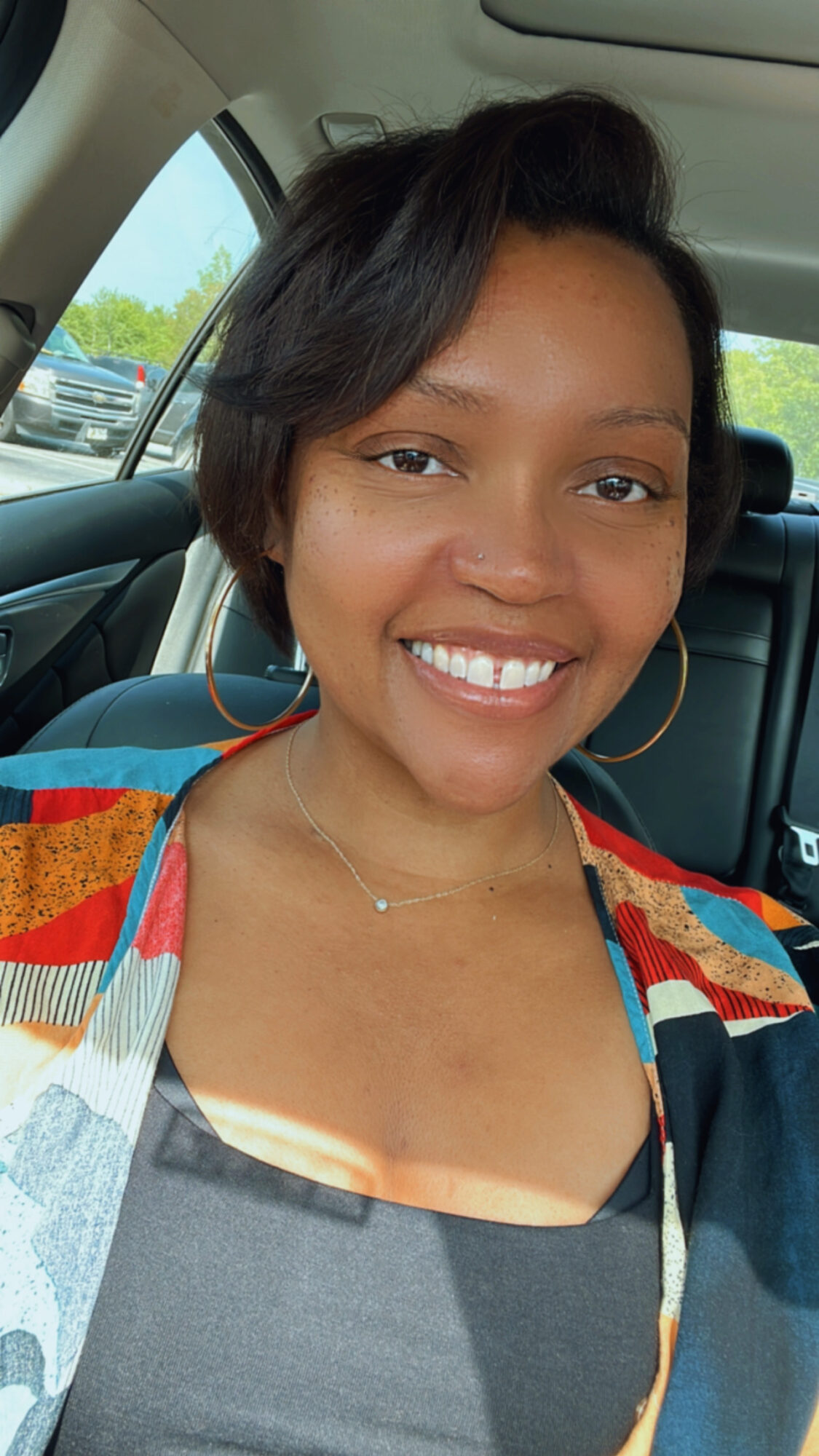 Smiling woman with short dark hair, wearing hoop earrings and a colorful top, sitting in a car.