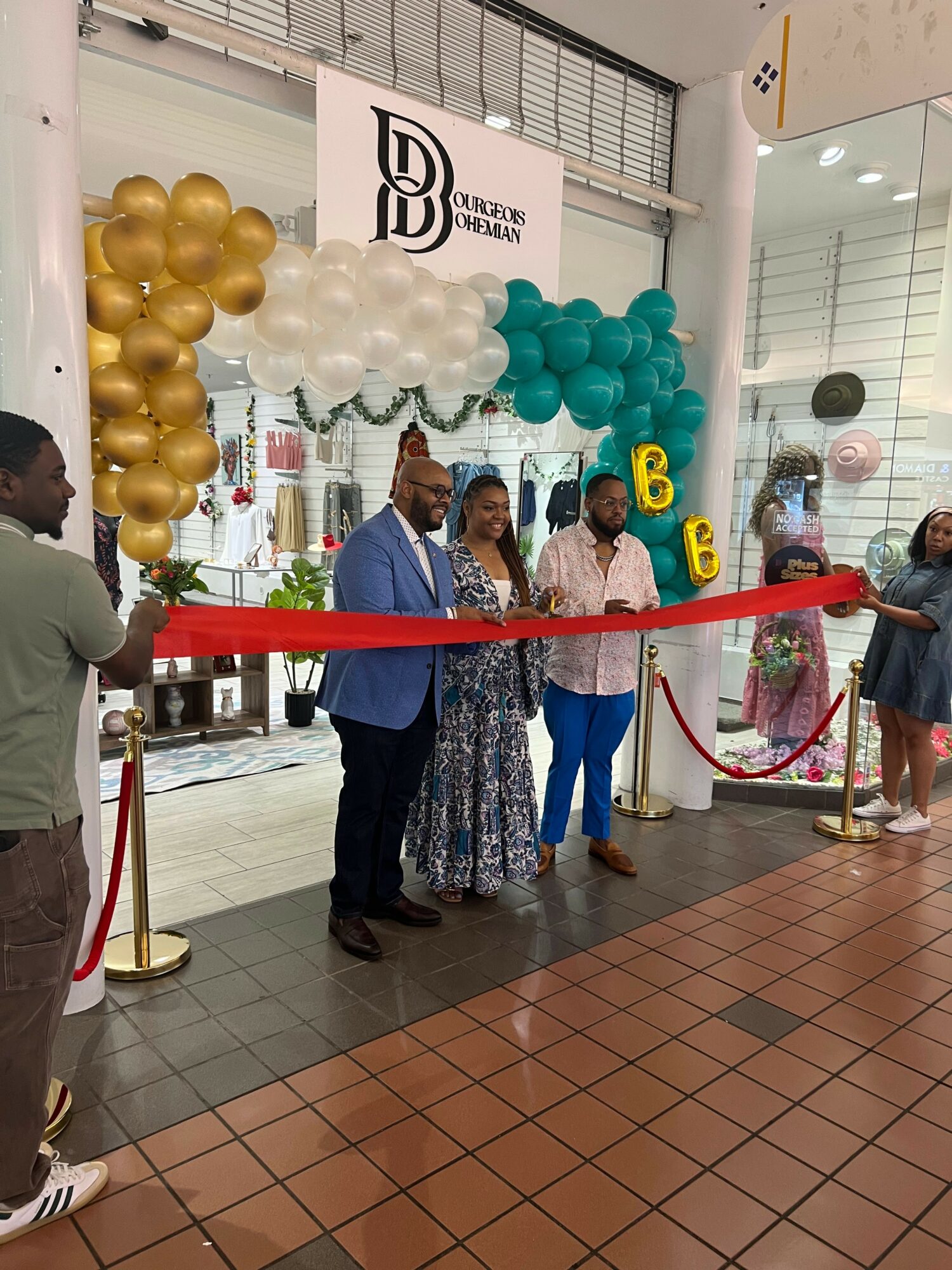 People standing in front of a ribbon at a store opening, decorated with balloons and a sign reading 'BD'.