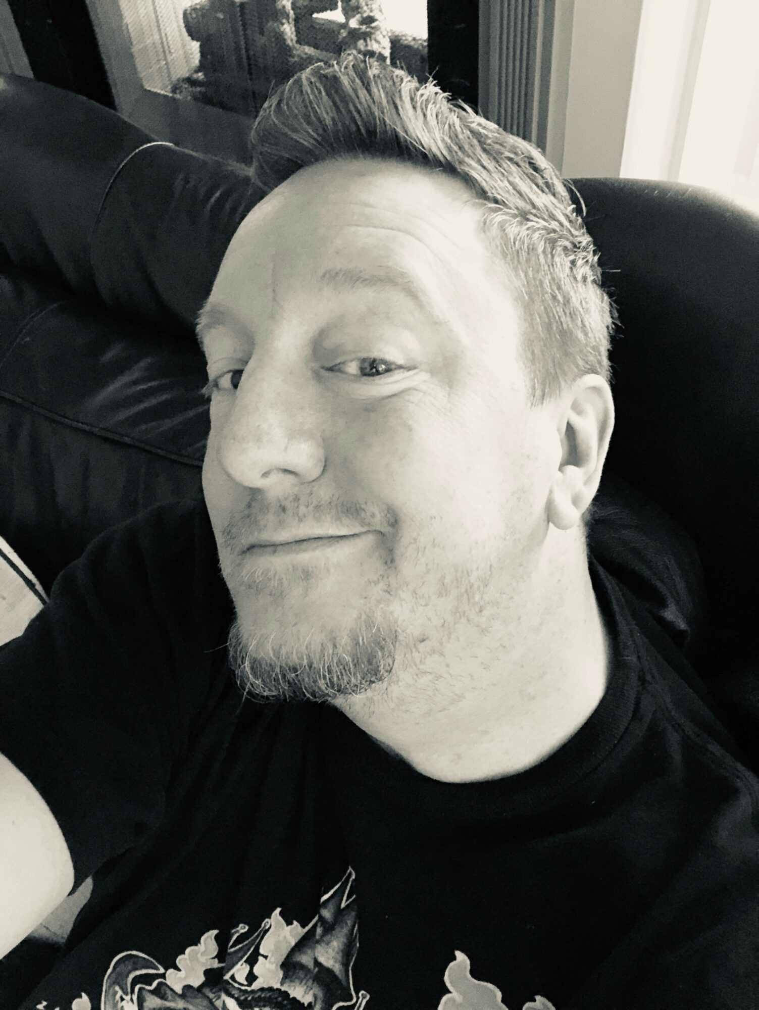 Black and white close-up of a smiling man with short hair and a beard, lying on a couch.