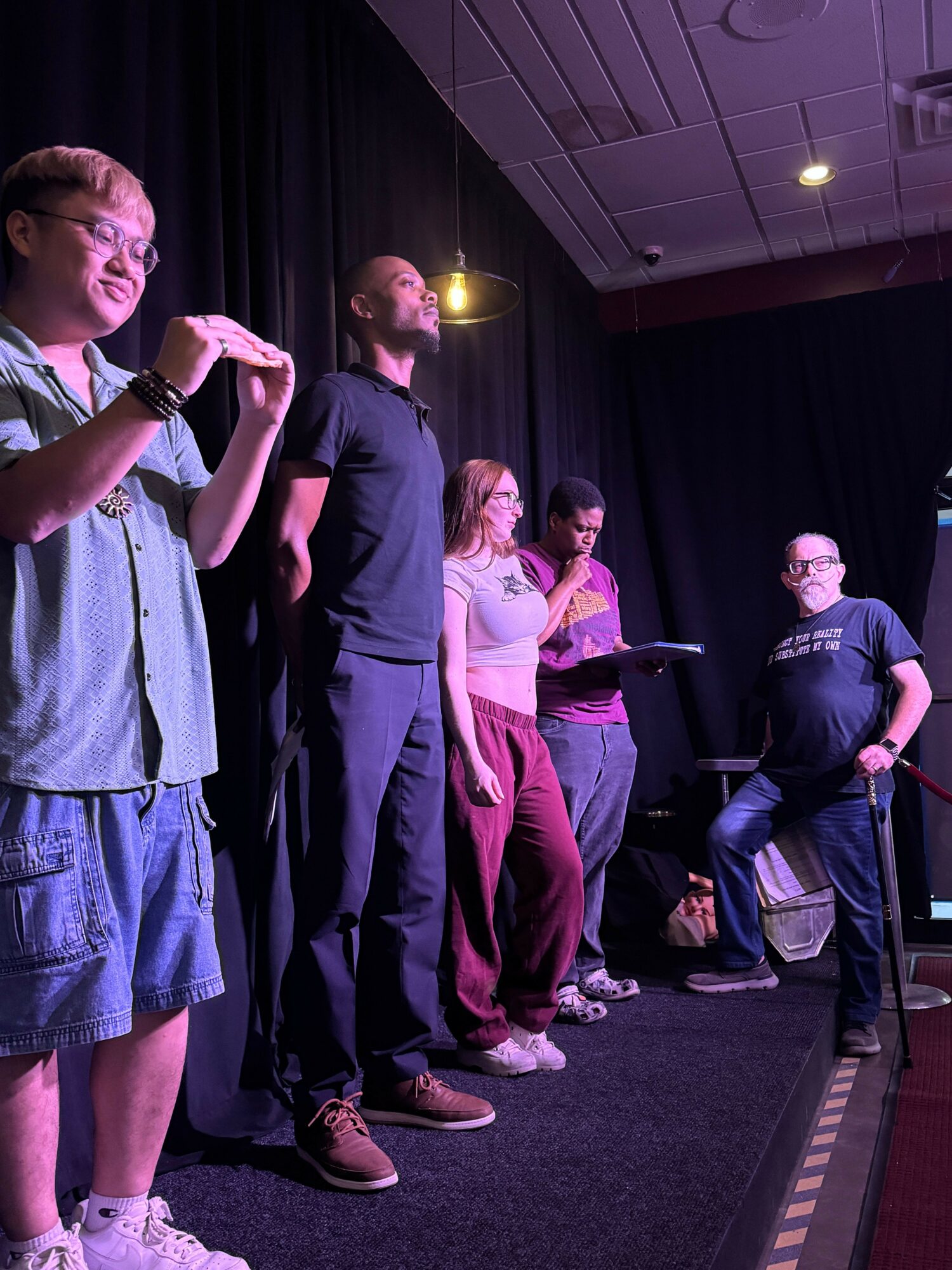 Group of people standing on stage, some talking, in a dimly lit room with black curtains and a dark floor.