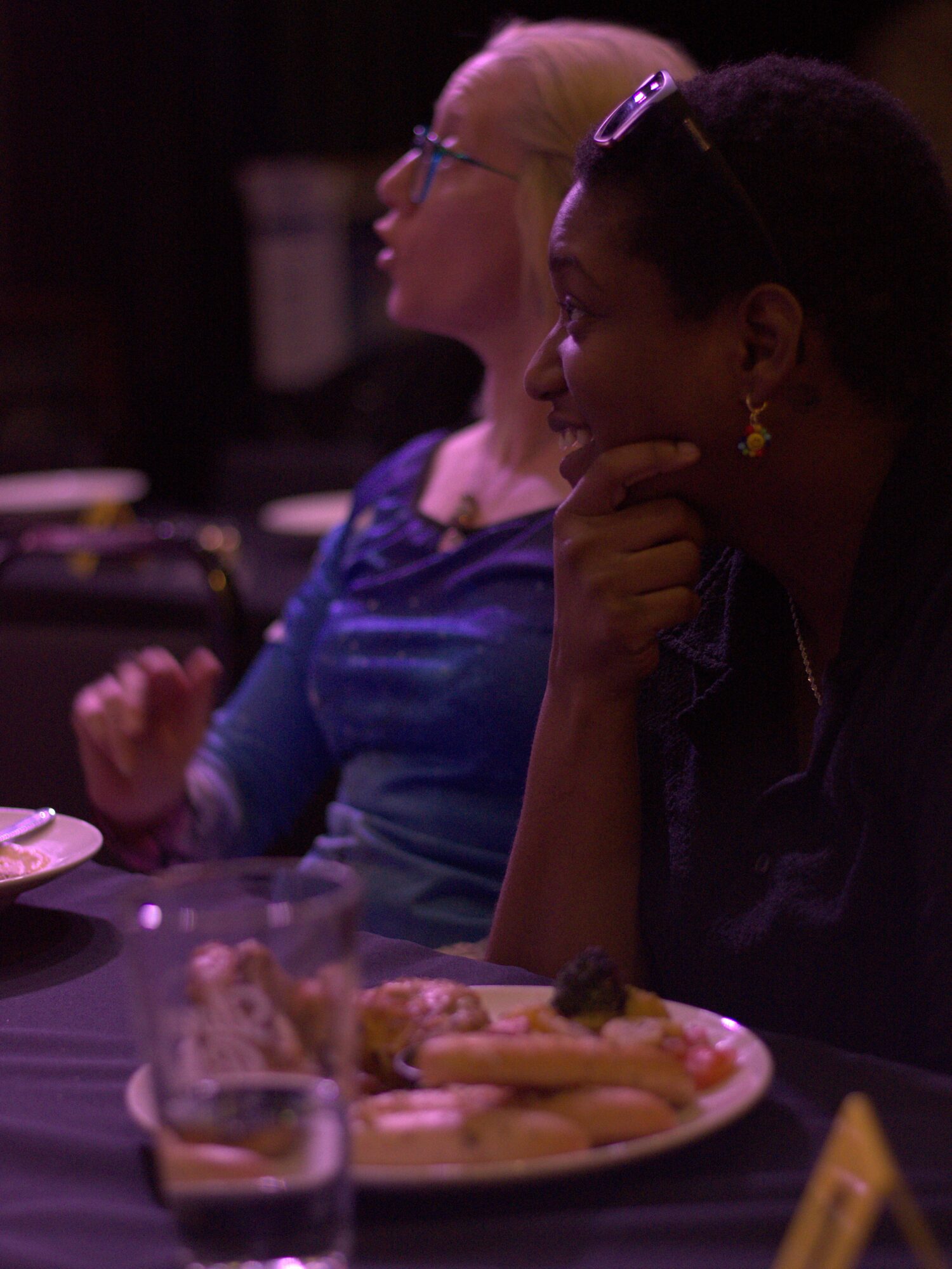Two women sit at a table with food and drinks, one with glasses and the other with a hand on her chin.