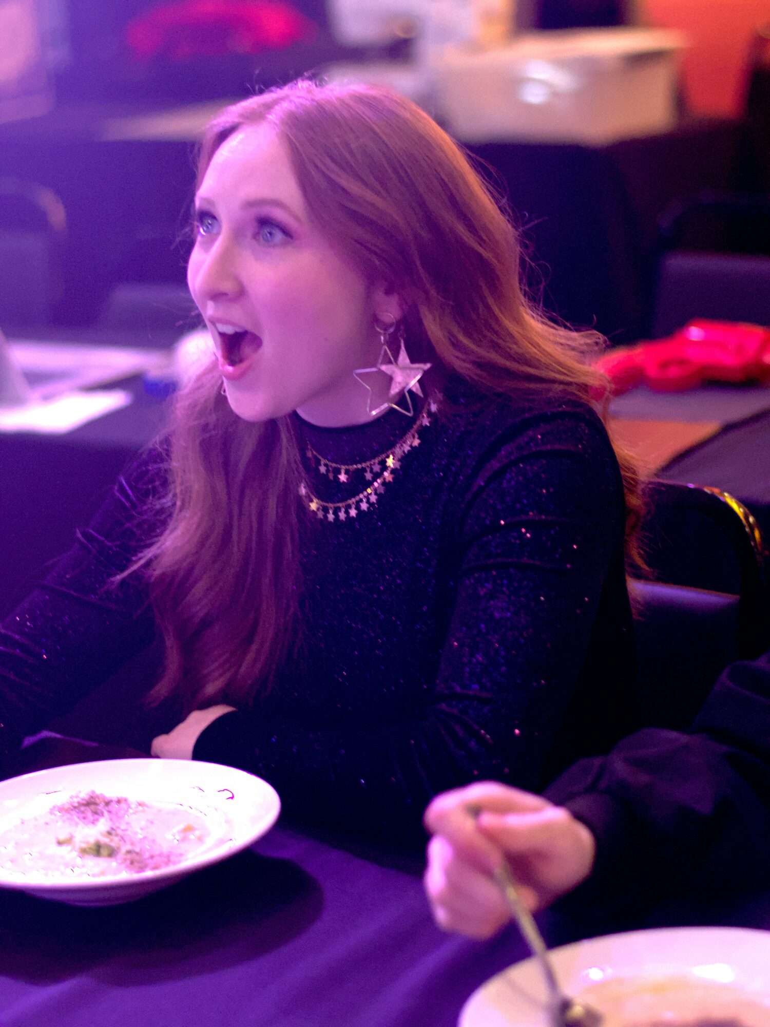 Young woman with long hair, wearing star-shaped earrings and a black top, appears surprised or excited at a social event.