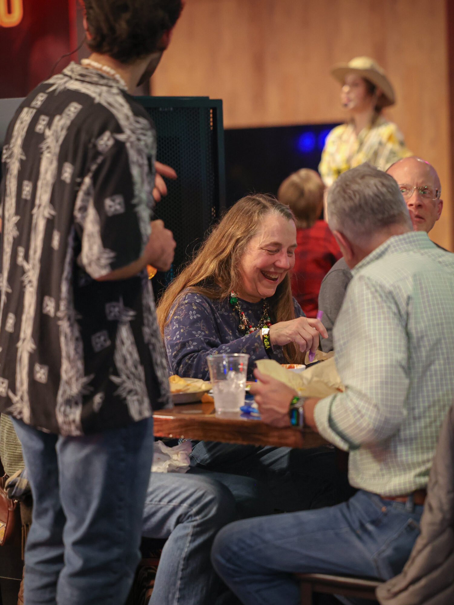 People gathered around a table, smiling and talking, with a woman in the background wearing a hat.