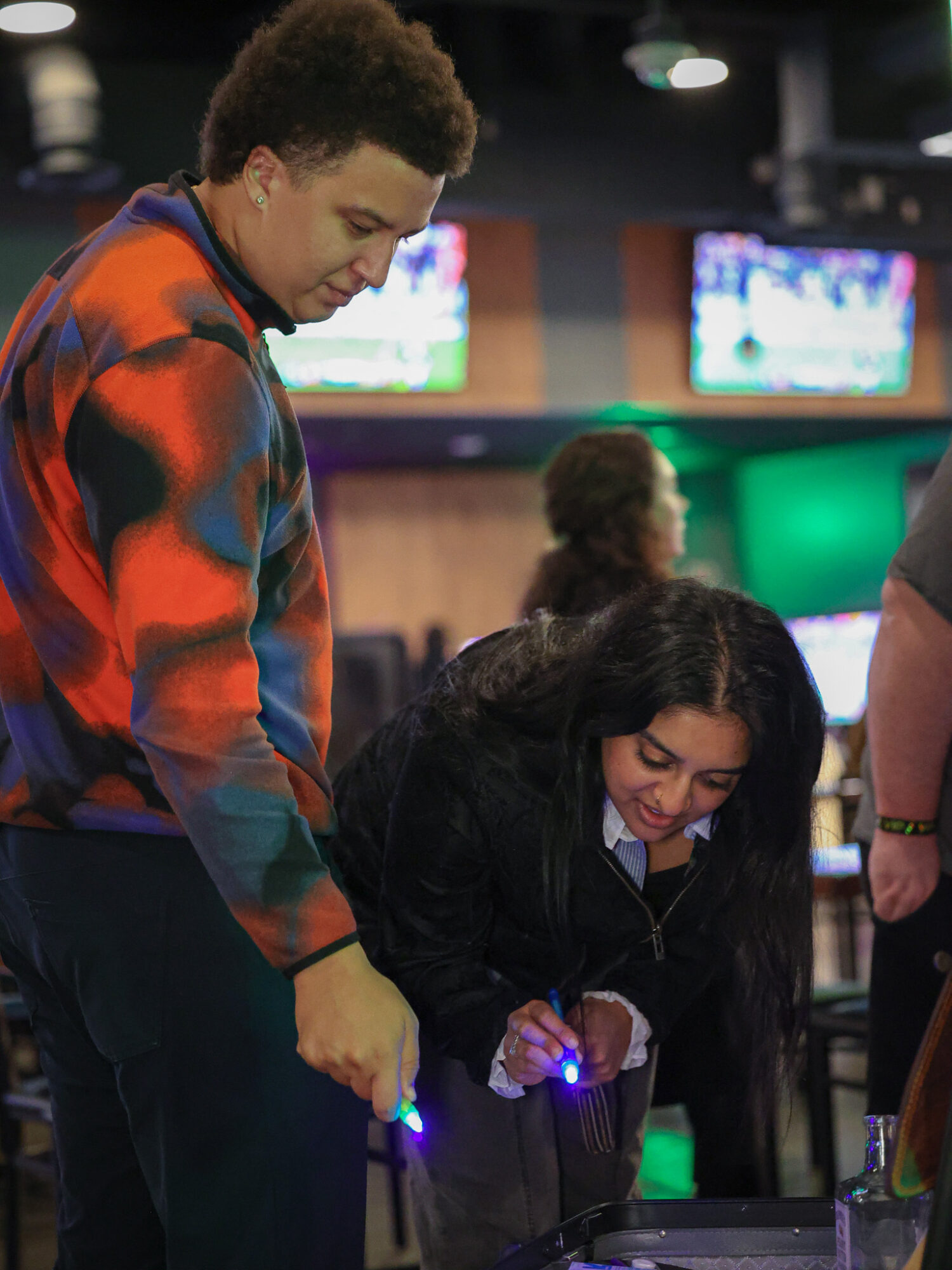 Two people, one standing and one sitting, in a gaming or entertainment venue with screens in the background.