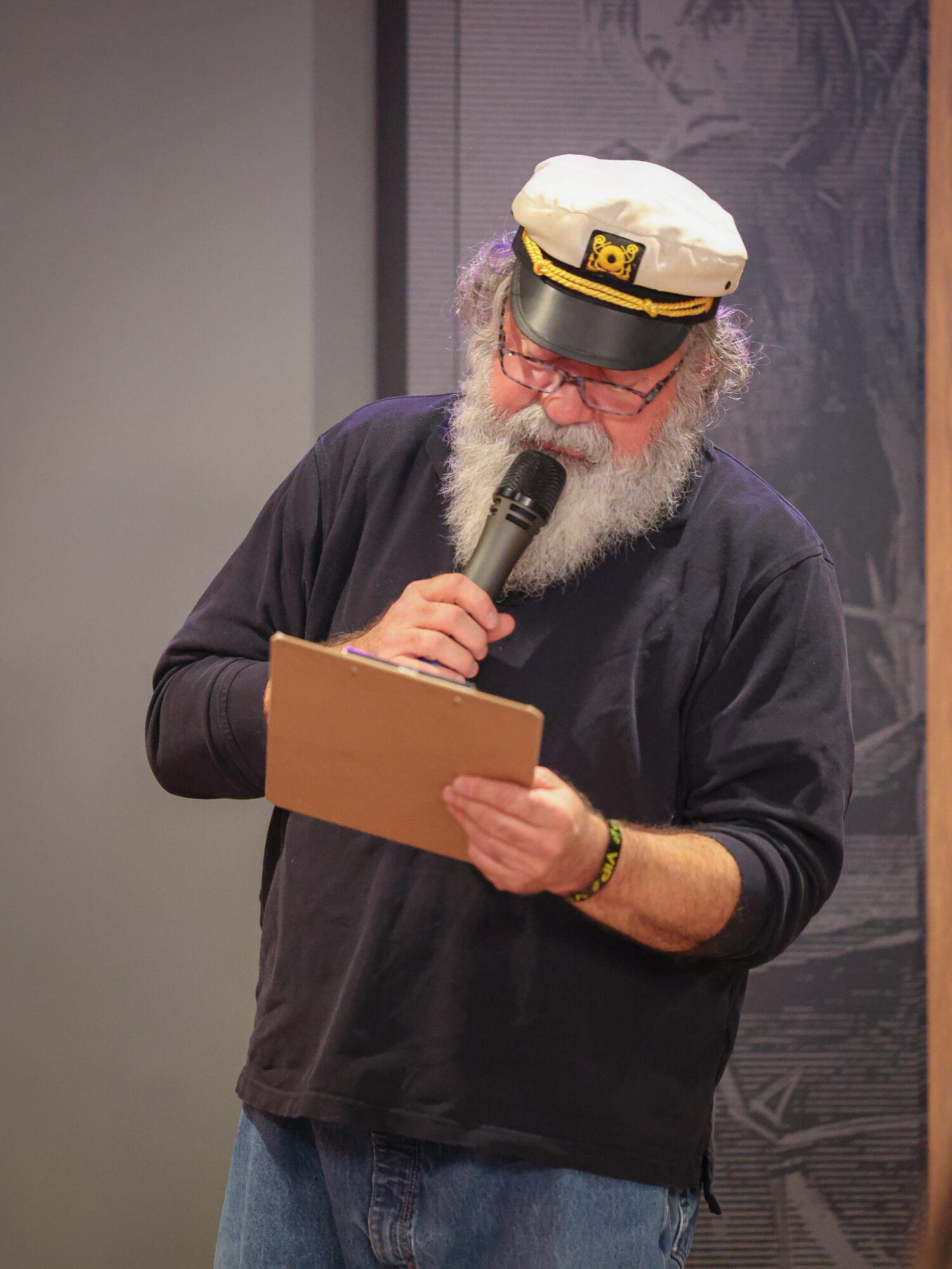 Older man with a white beard and glasses, wearing a captain's hat, reading from a clipboard and speaking into a microphone.