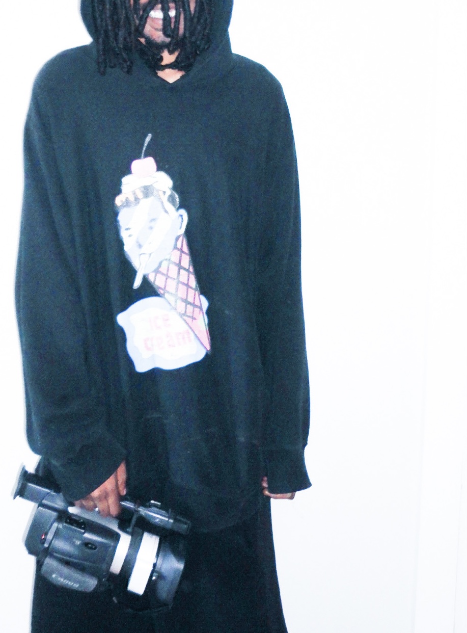 Person with dreadlocks wearing a black hoodie with ice cream cone graphic, holding a camera, against a white background.