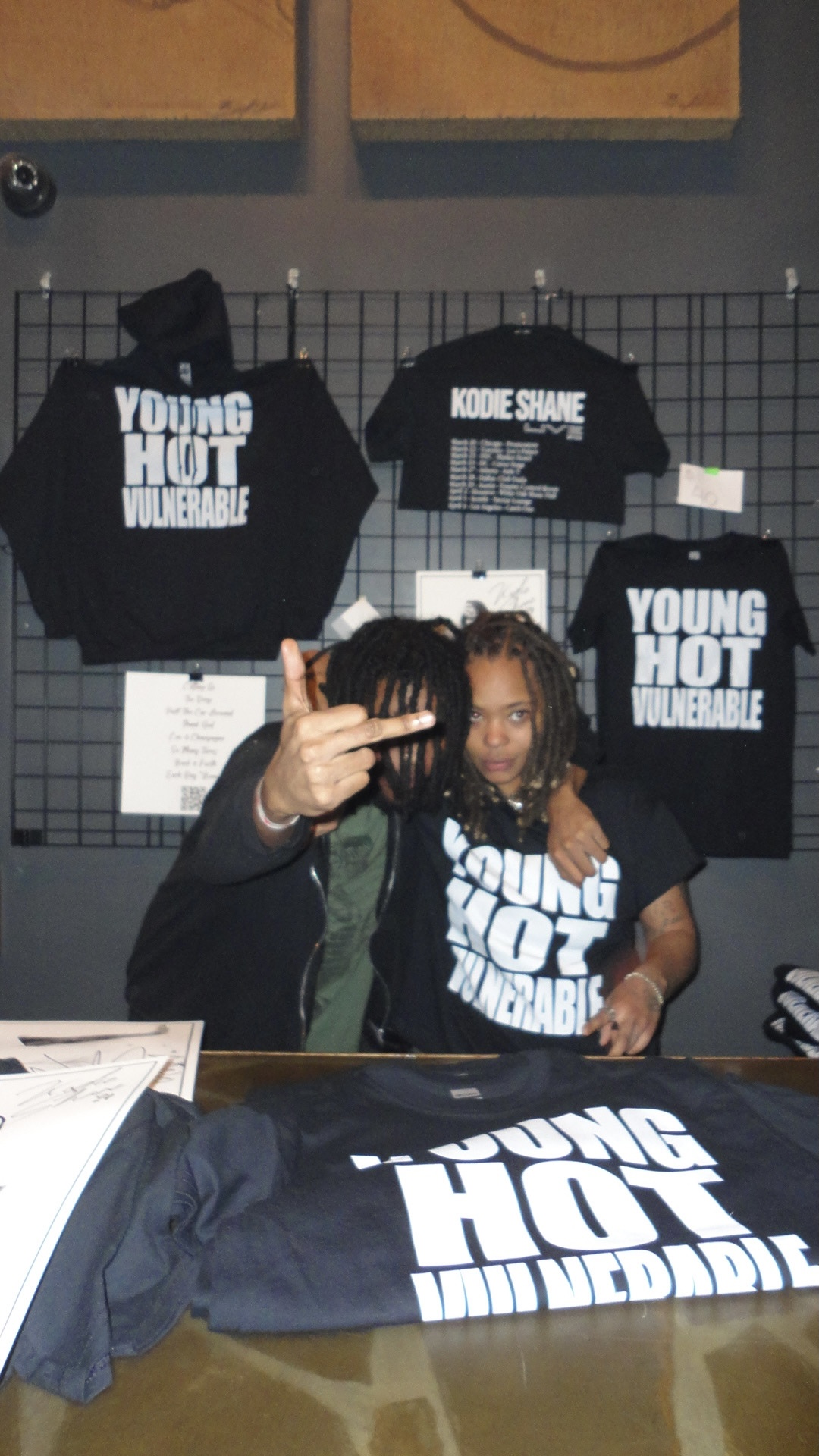 Two children sit at a table with black T-shirts and hats displaying text, behind a wire grid display, with more shirts hanging.