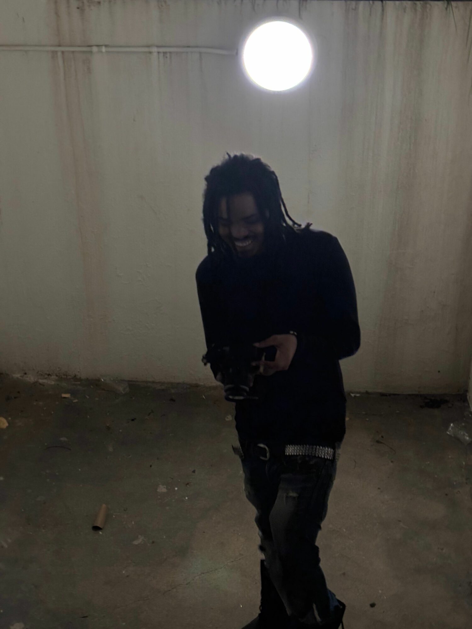 Person standing in a room with a curtain and a bright light, holding a phone, facing camera, with dark clothing.