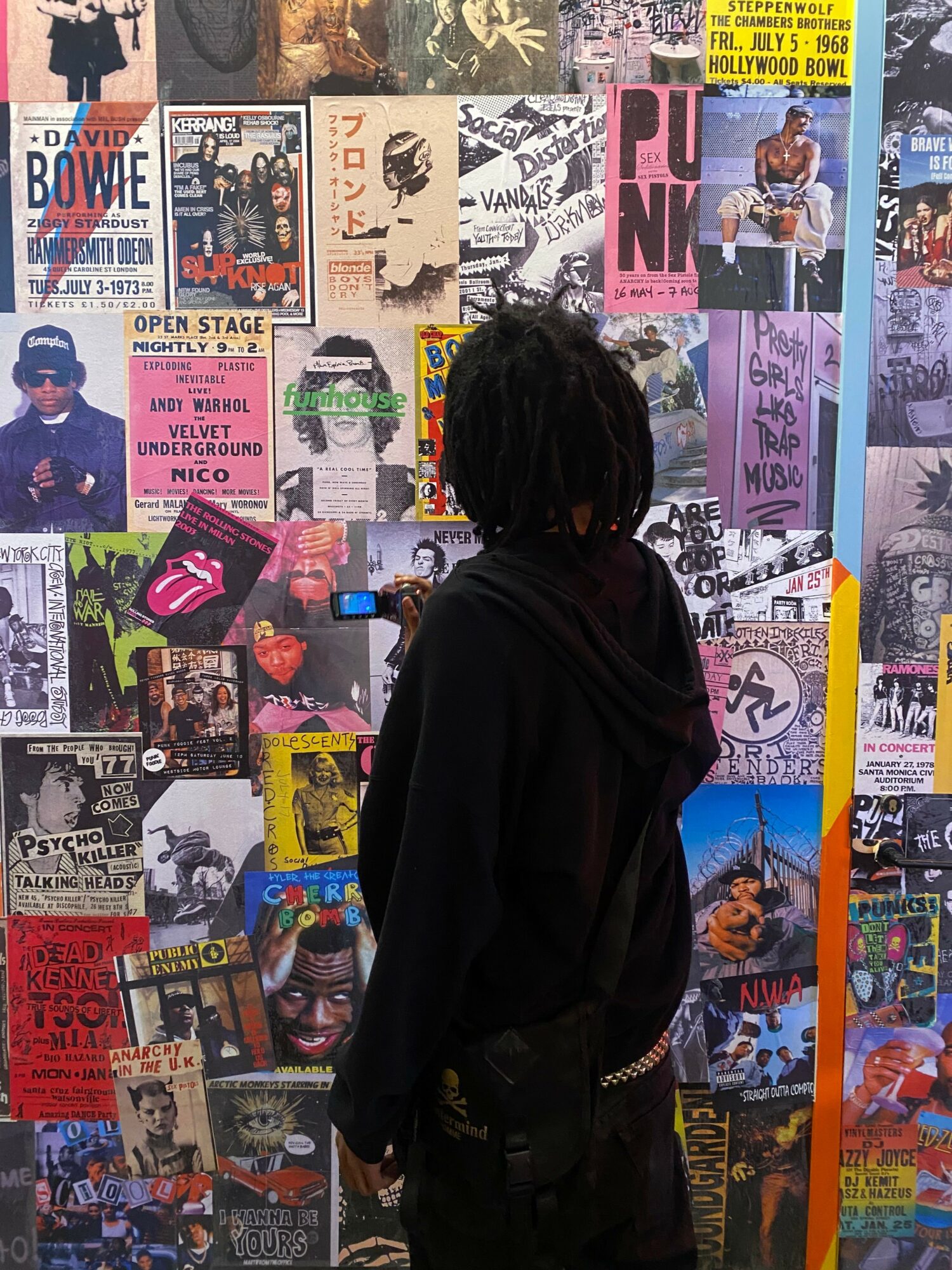 Person with black hair and black clothing looking at a colorful collage of posters and images on a wall.