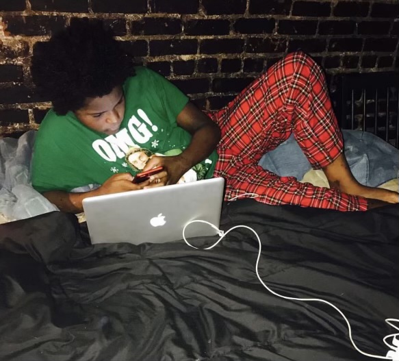 Person lying on bed with a laptop, phone, and charging cable, against a brick wall background.