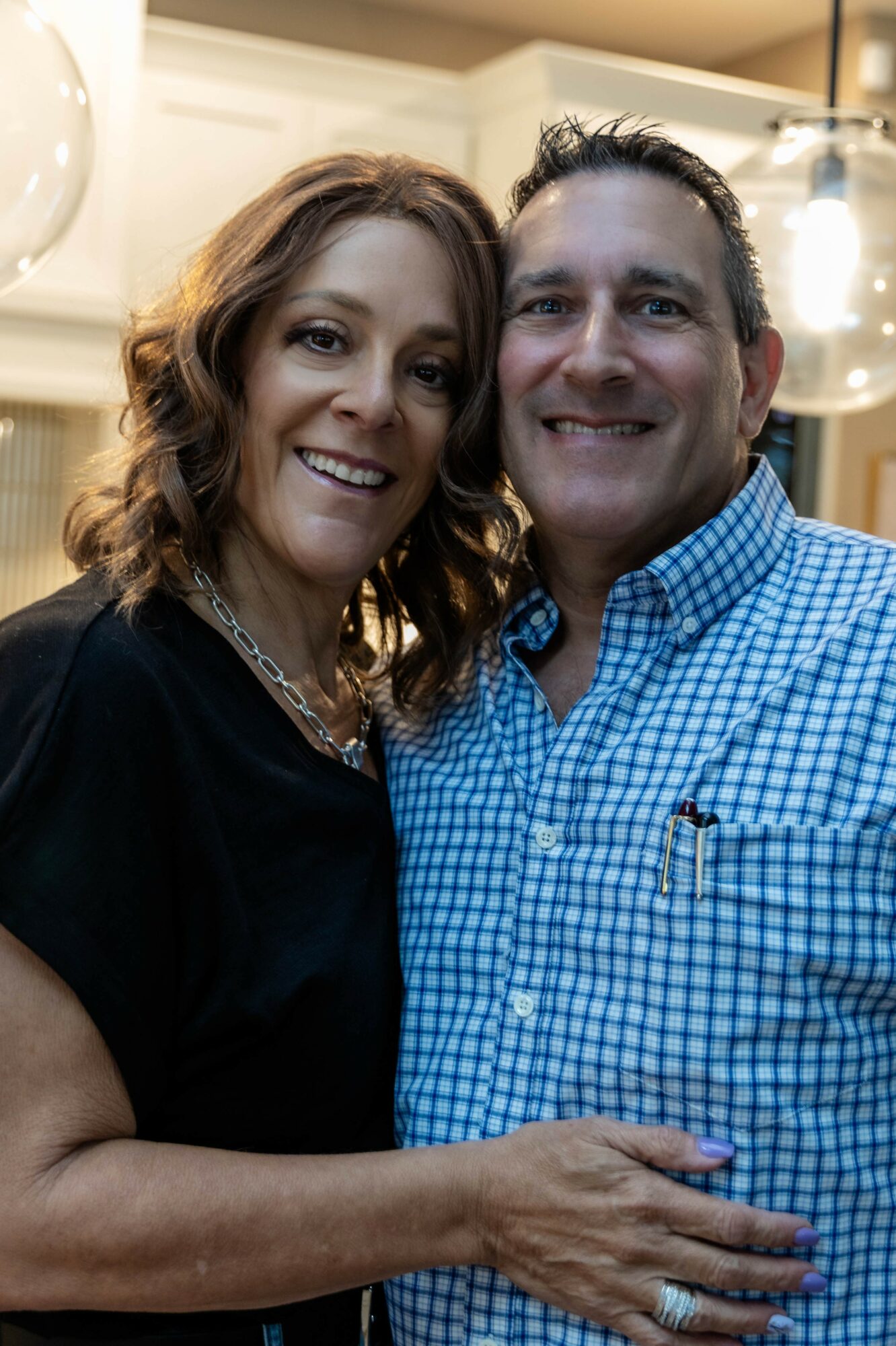 A smiling woman with wavy hair and a man in a blue checkered shirt pose together indoors.