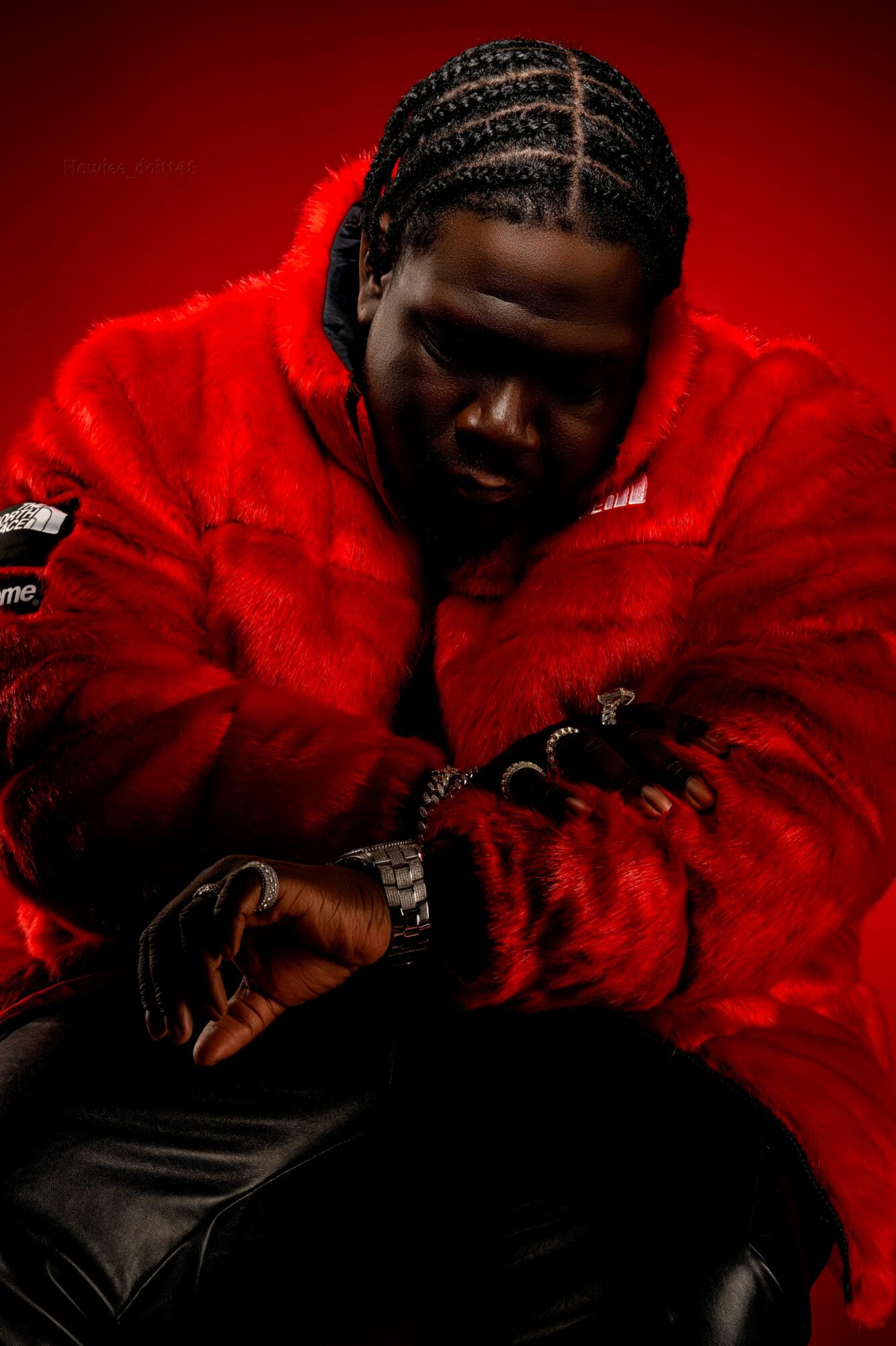 Person with braided hair wearing a red fur jacket, sitting with arms crossed against a red background.
