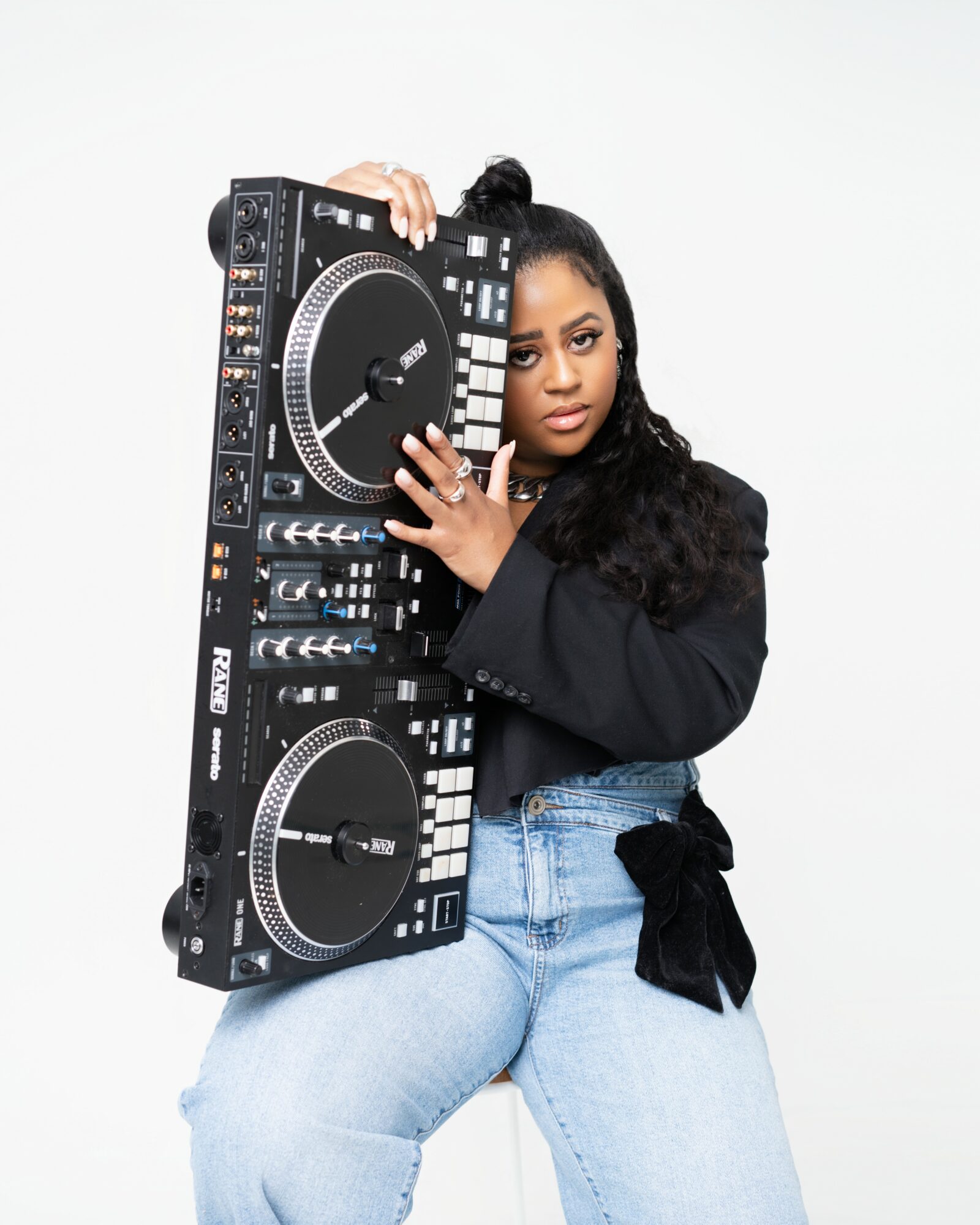 Woman holding DJ controller, looking at camera, wearing black jacket and light blue jeans, against a white background.