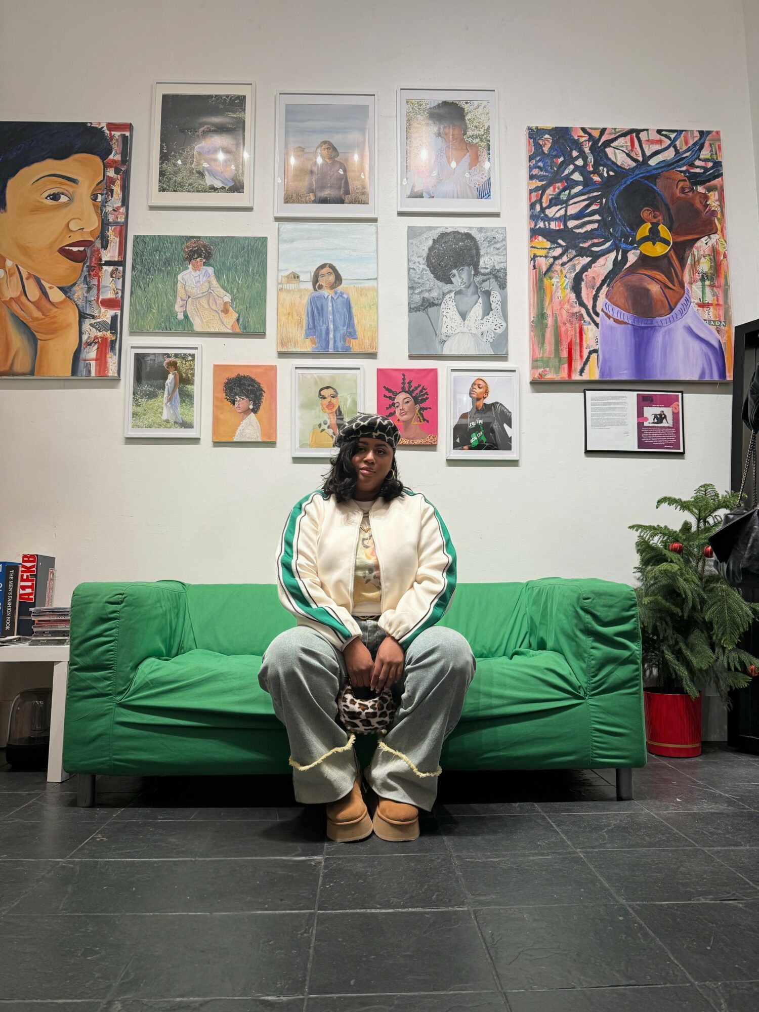 Person sitting on a green sofa in front of a wall with colorful artwork, wearing a white jacket and beige pants.