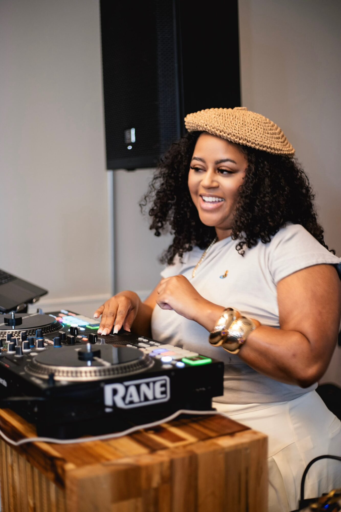 Woman with curly hair wearing a hat and white dress, smiling while DJing with equipment on a wooden table.