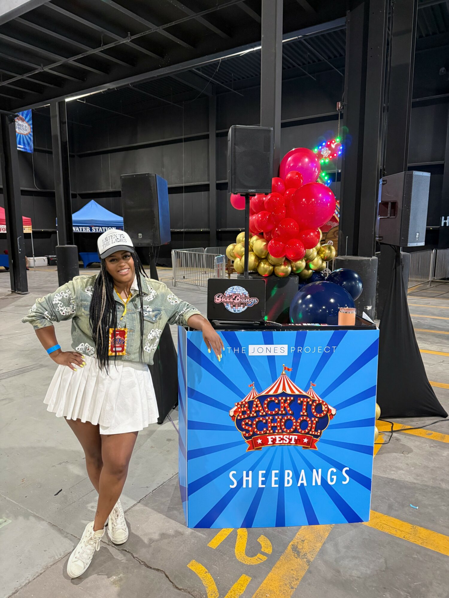 Young woman in a white skirt, patterned jacket, and white hat standing next to a colorful balloon display and a sign at an indoor event.