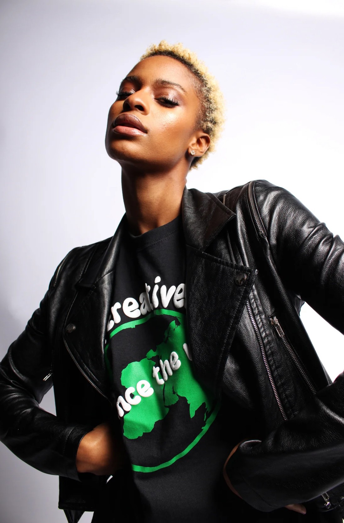Young woman with short blonde hair wearing a black leather jacket and graphic t-shirt, posing confidently against a plain background.