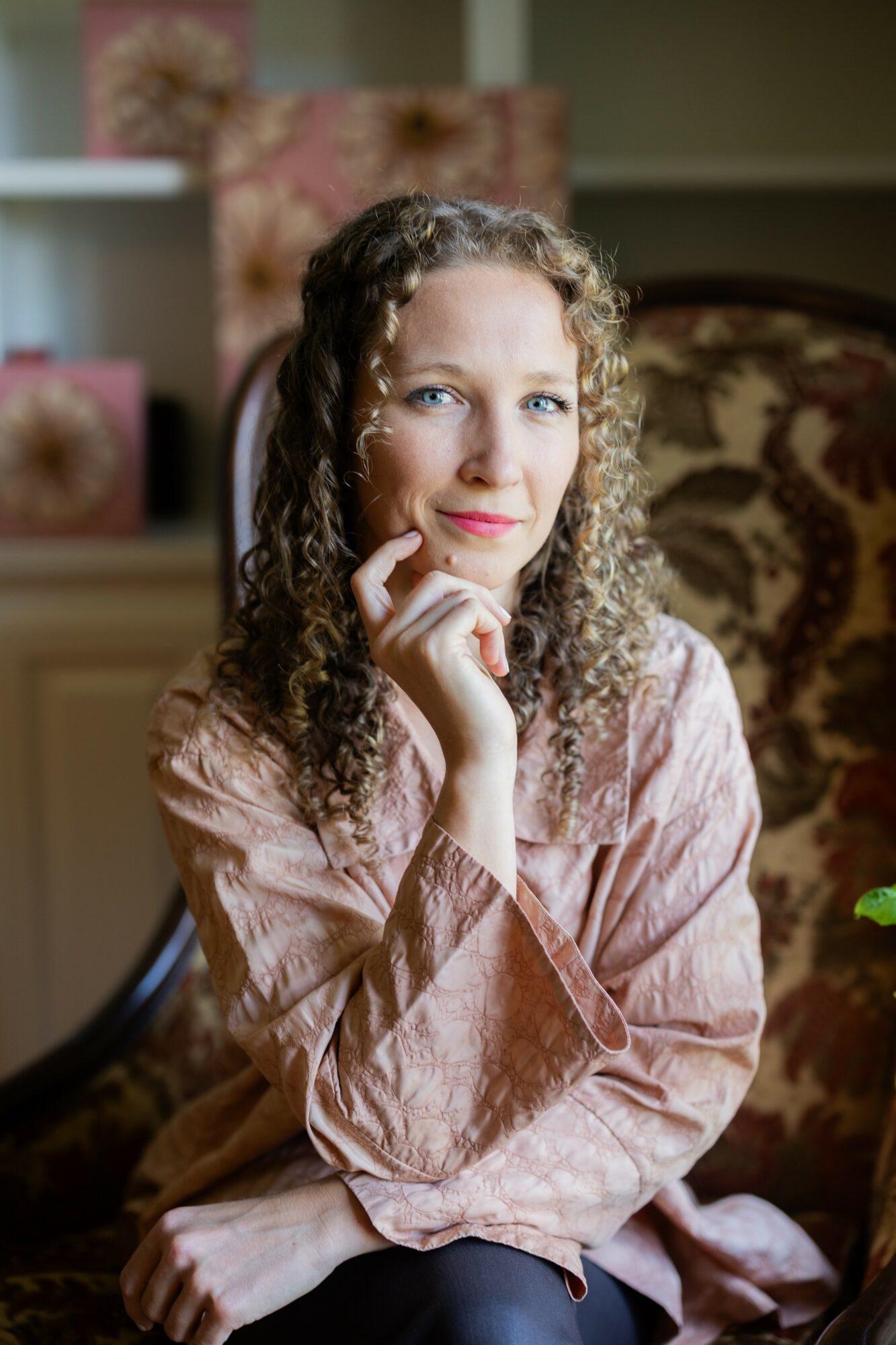 Young woman with curly hair sitting on a patterned armchair, smiling, hand near chin, indoors.