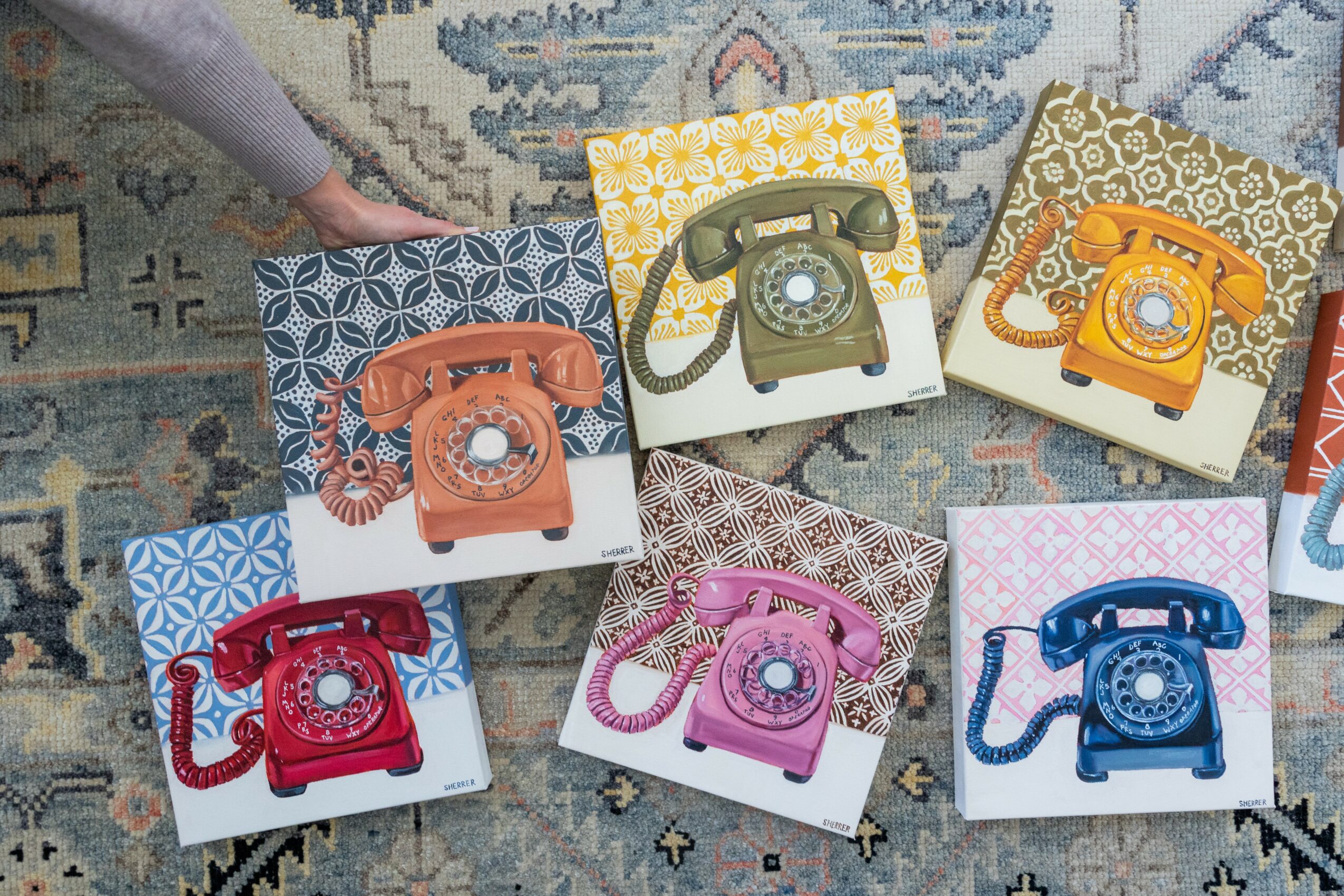 Six colorful vintage rotary phones on patterned boxes, arranged on a rug, with a person's hand visible in the top left corner.