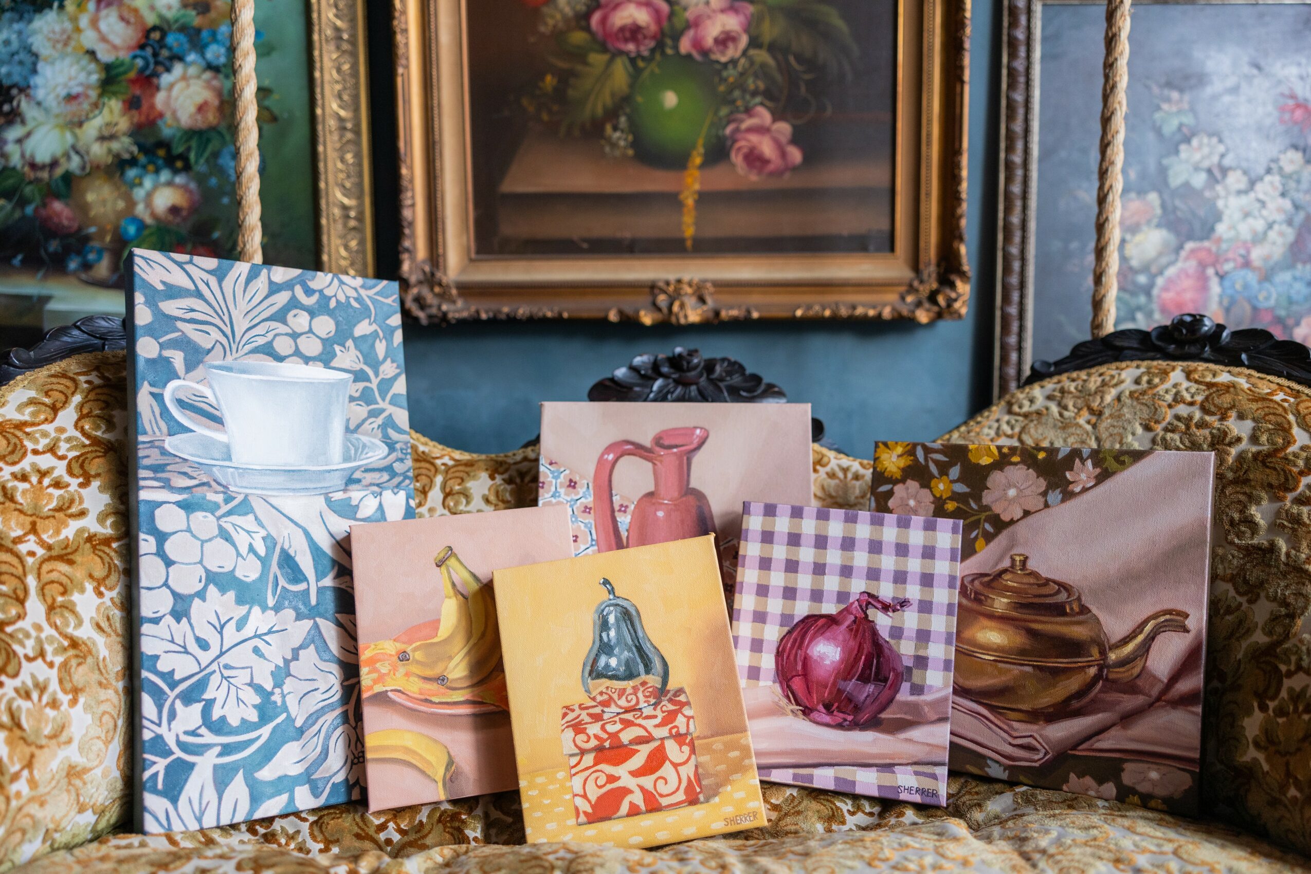 Collection of greeting cards with floral, teapot, and fruit designs, arranged on a sofa with patterned cushions, framed paintings in background.