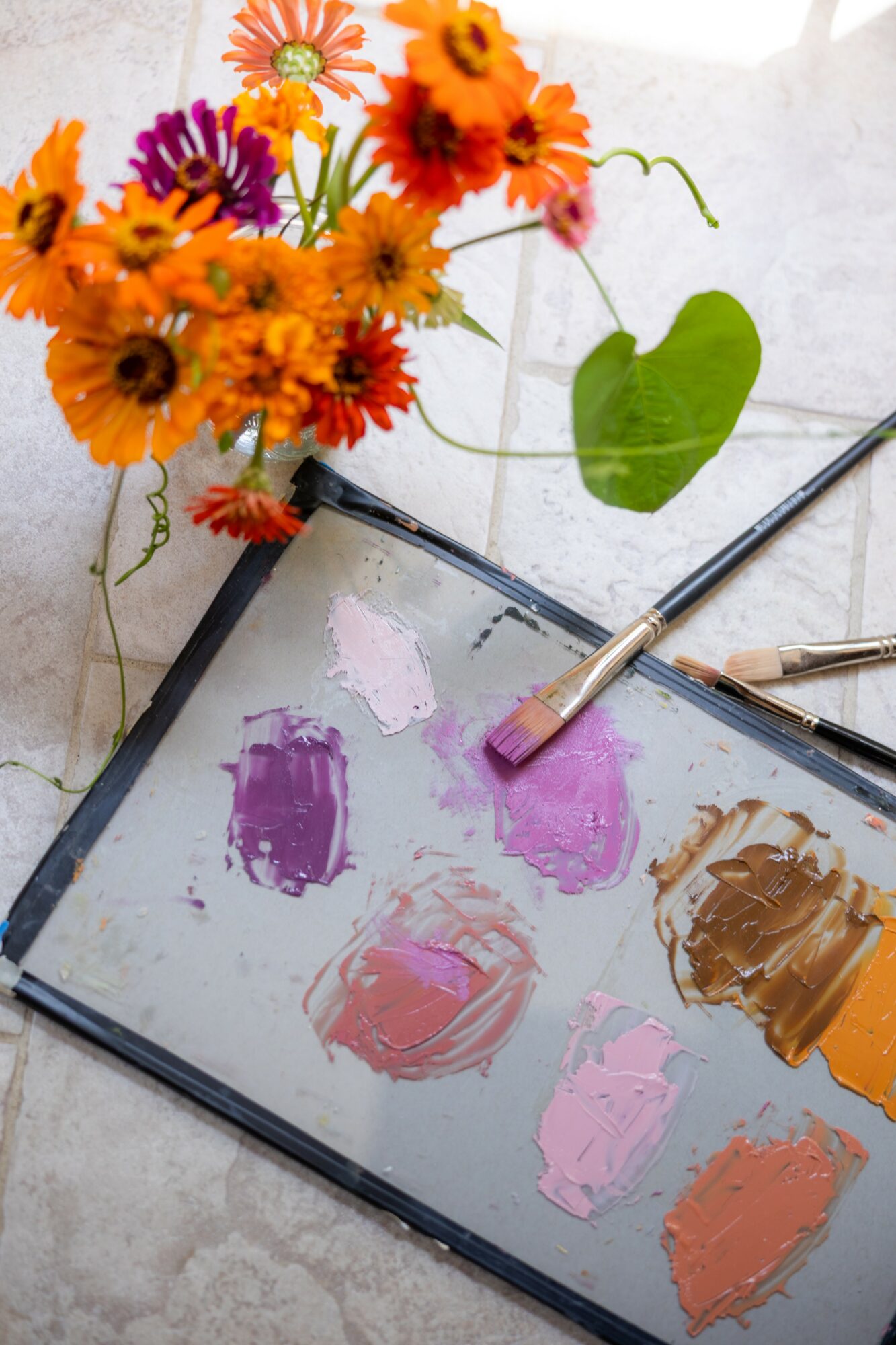 Paint palette with pink, purple, and brown paint, with a bouquet of orange, purple, and yellow flowers nearby on a white surface.
