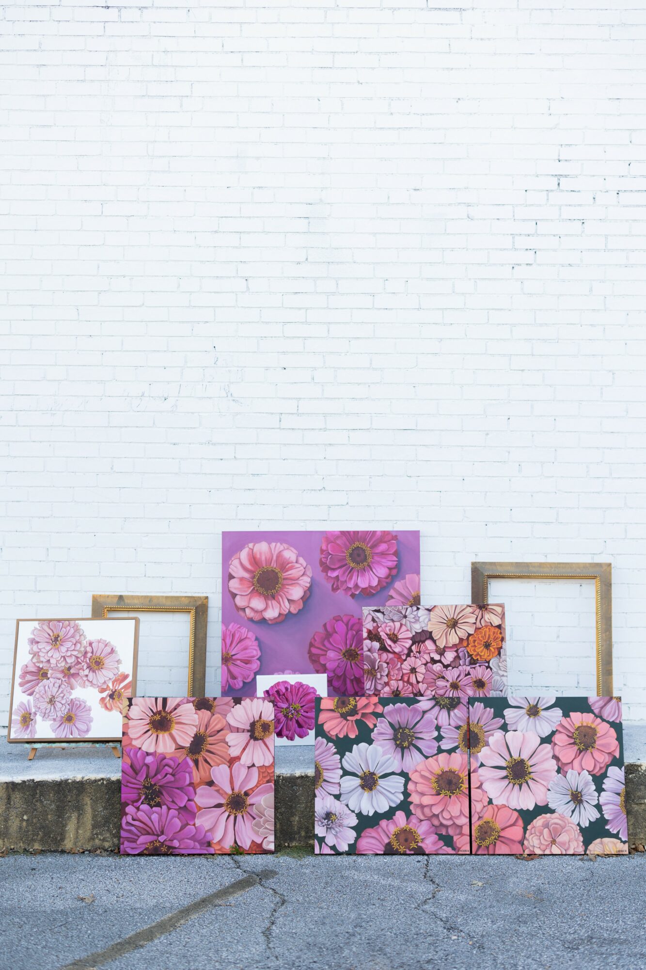 Collection of floral paintings with pink and purple flowers, arranged against a white brick wall, with empty frames behind them.
