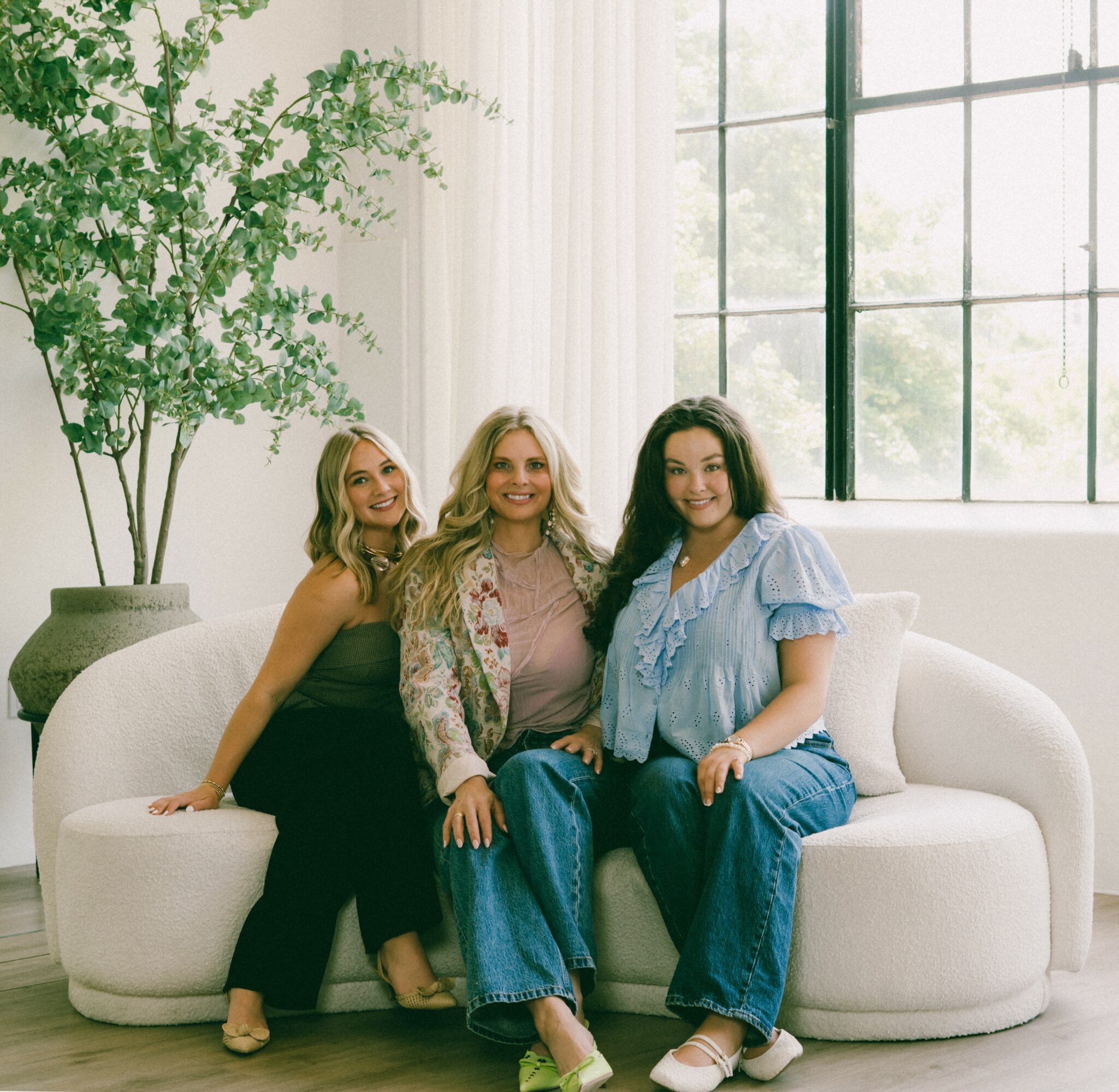 Three women sitting on a white sofa in a bright room with large windows and a potted plant.