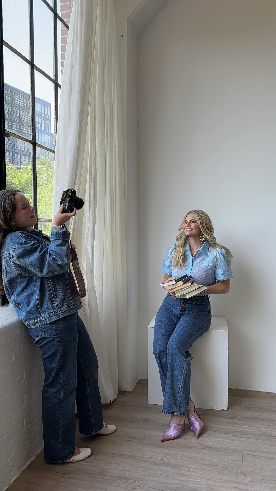 Two women near a large window with white curtains, one taking a photo of the other sitting on a stool, holding a book.