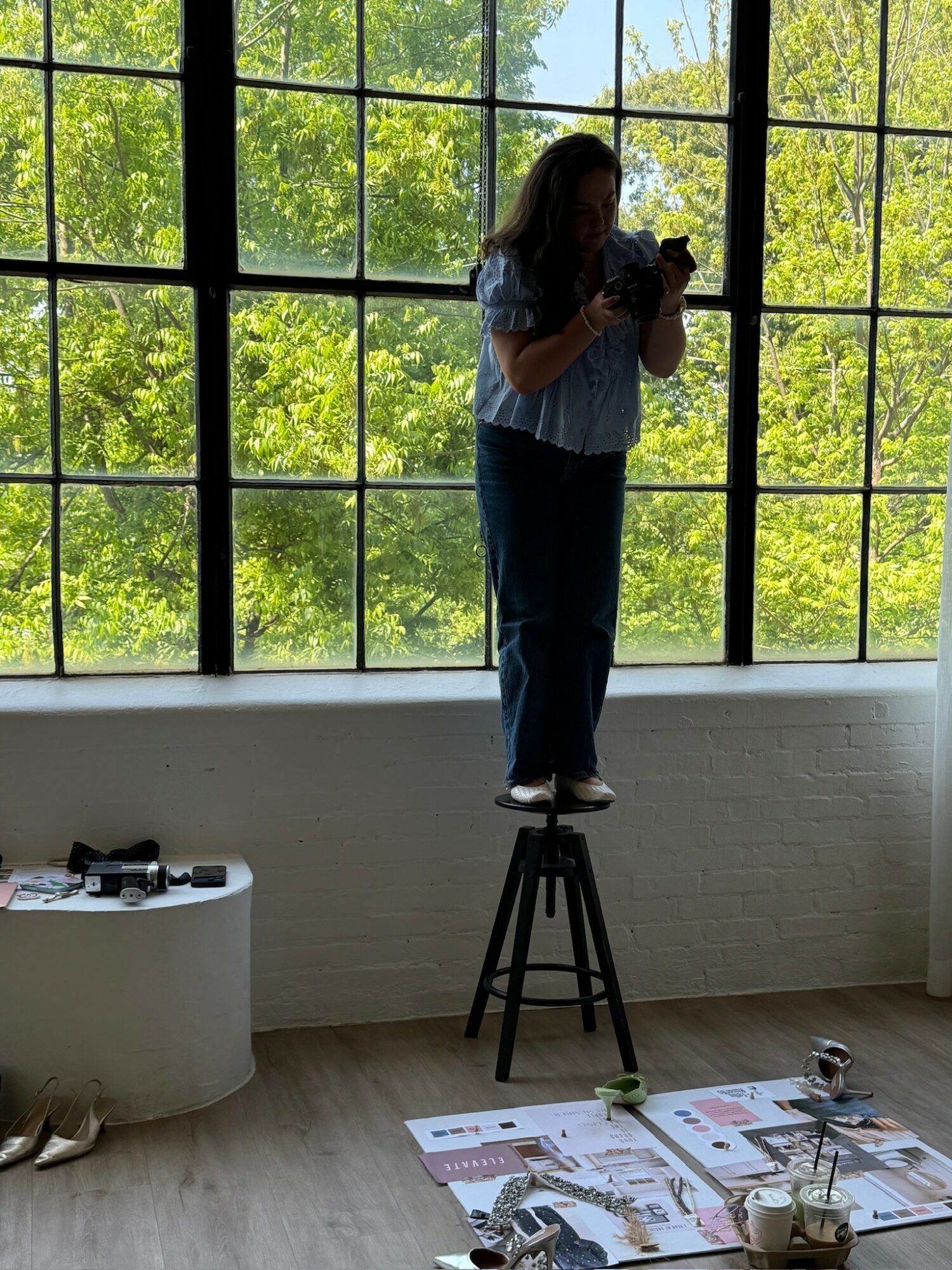 Person standing on a stool taking a photo indoors, large window with green trees outside, items on the floor and a table nearby.