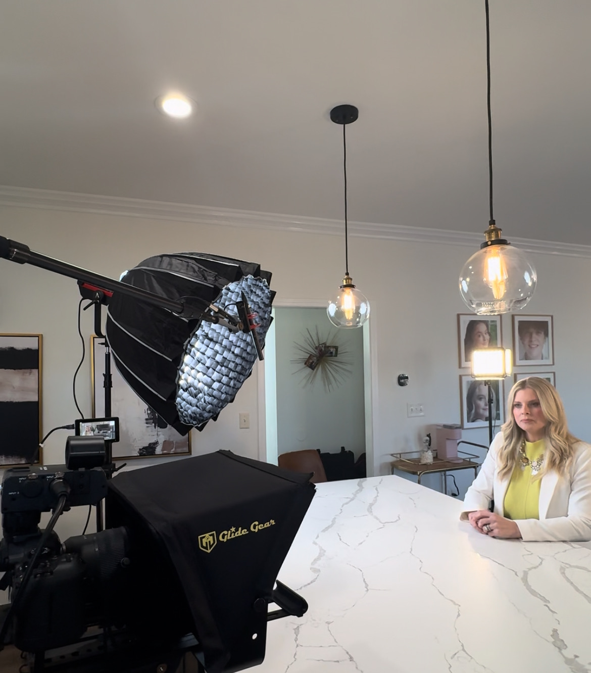 Woman sitting at a marble countertop in a well-lit room with hanging pendant lights and framed pictures on the wall.