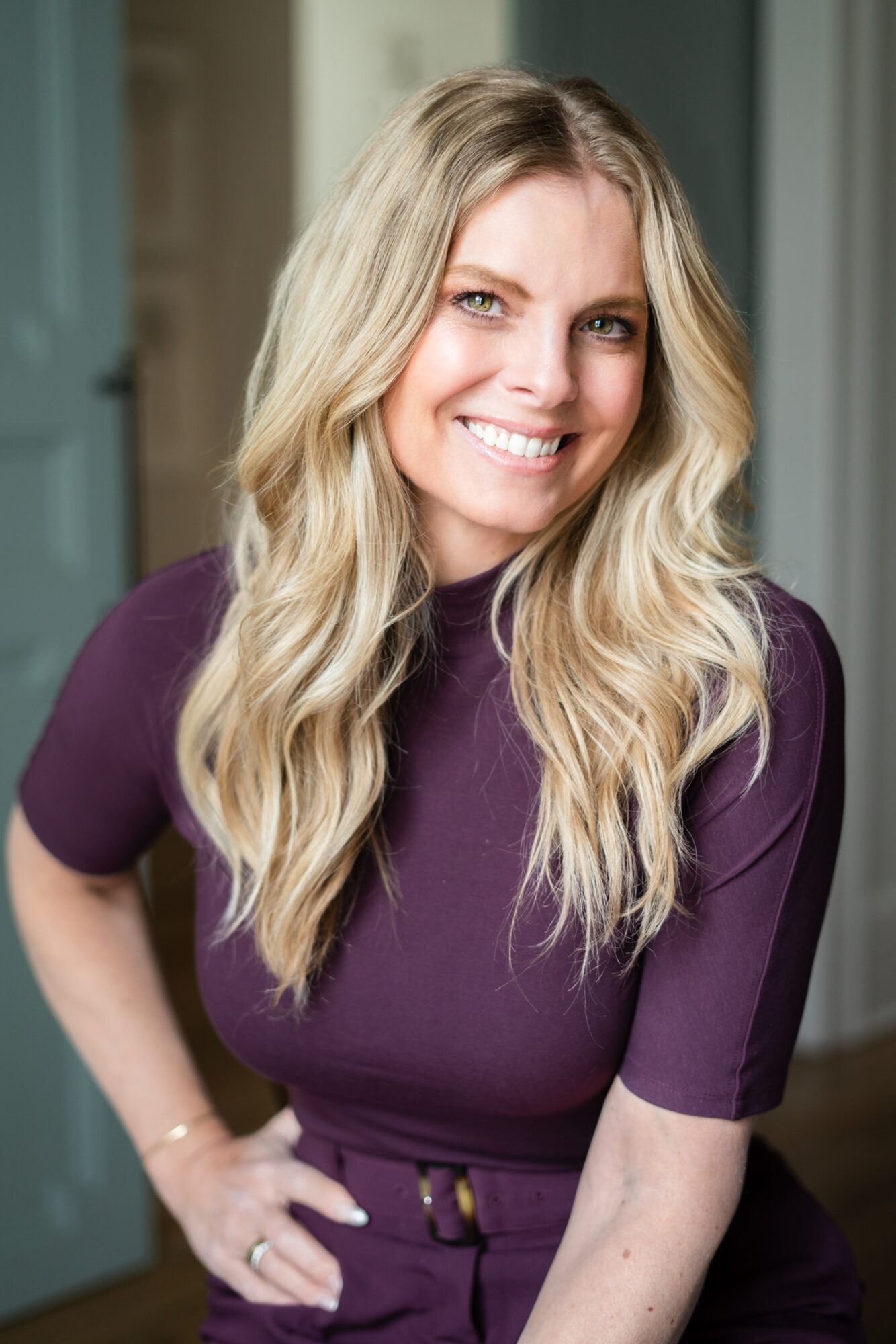 Woman with long blonde hair smiling, wearing a purple top, standing indoors with a blurred background.