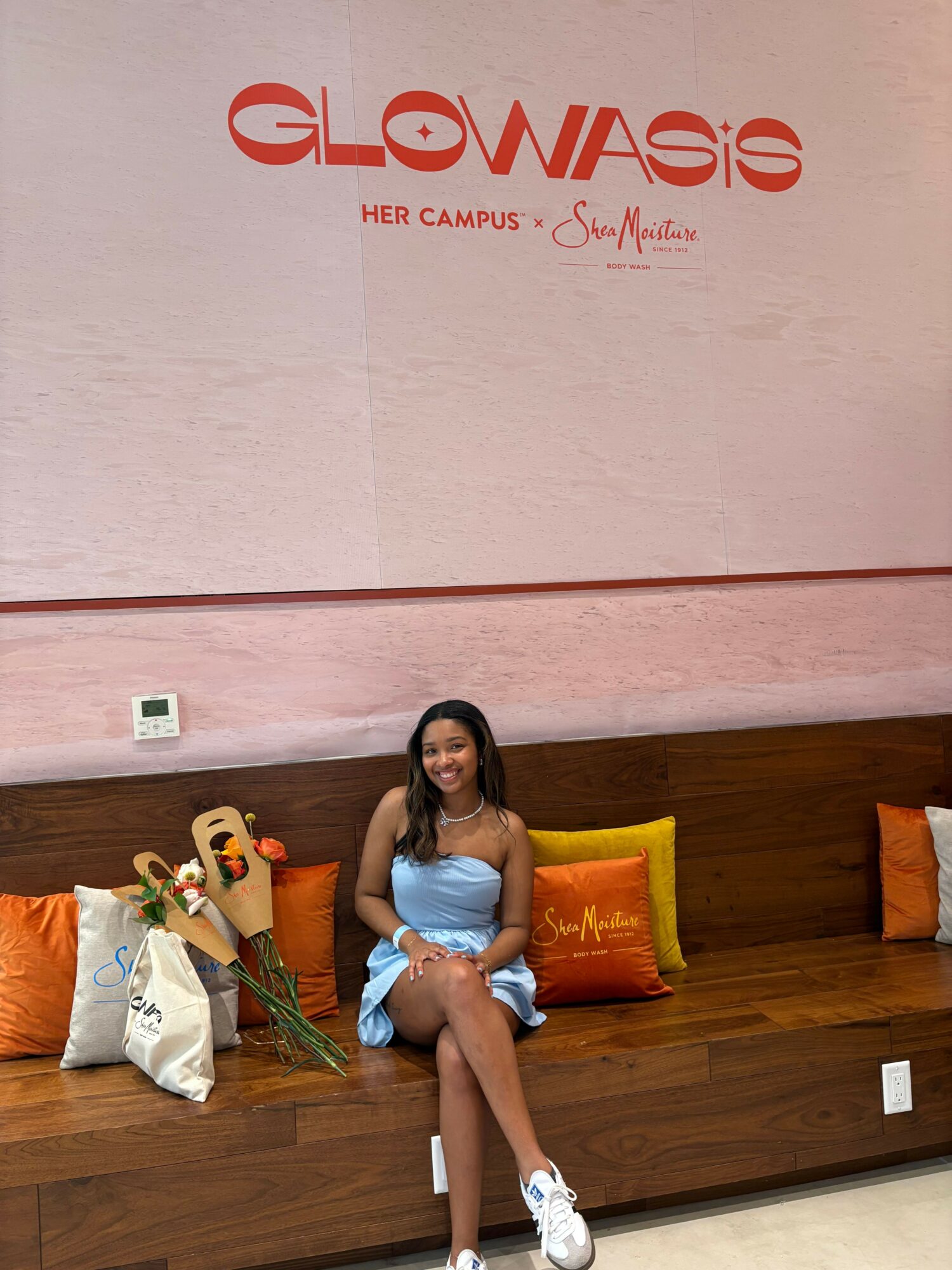 Woman sitting on a wooden bench with bags and flowers, wall with logo and text behind her, indoors.