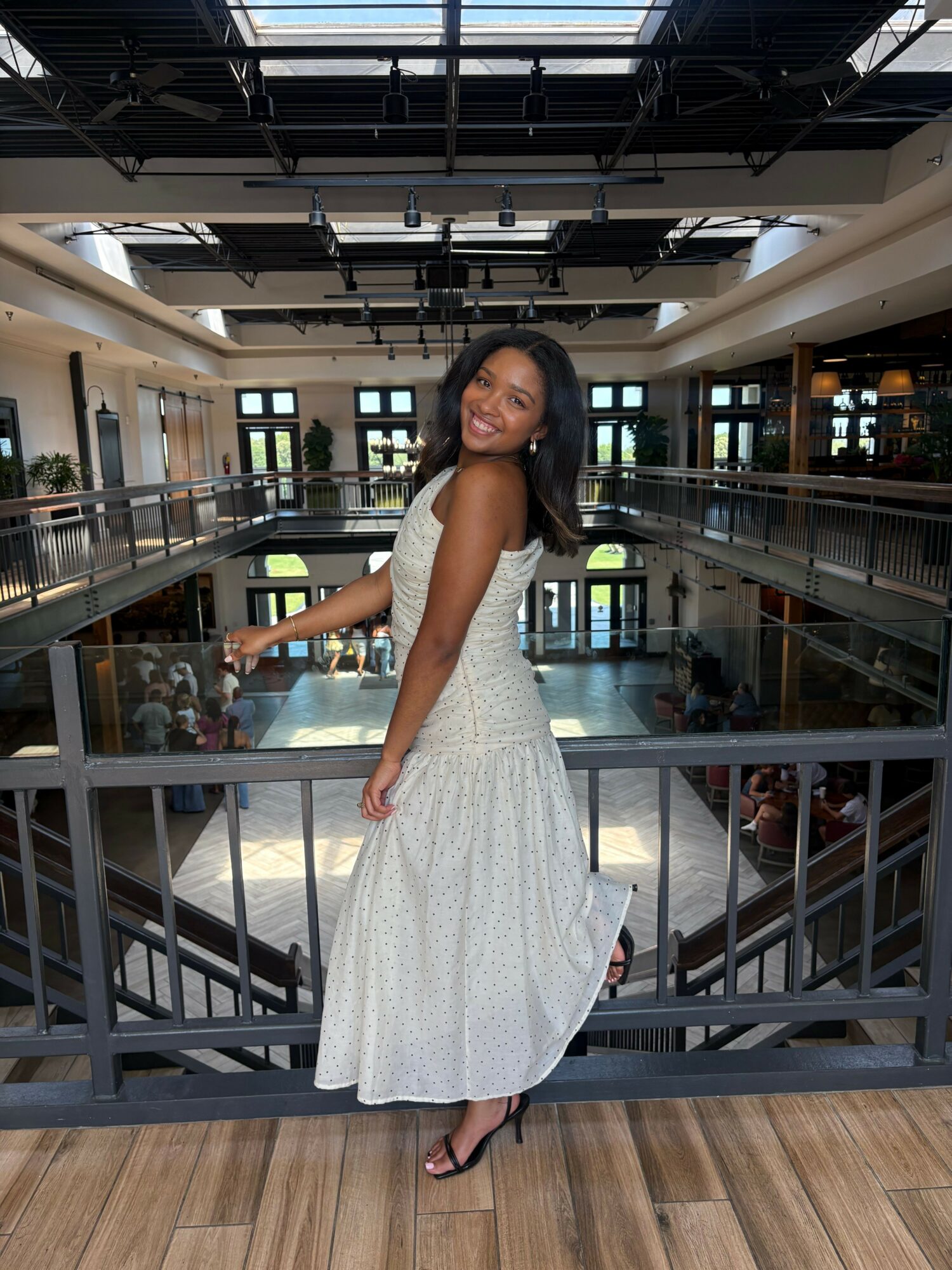 Woman in a white dress standing on a wooden floor, smiling, inside a modern building with glass and metal structures.