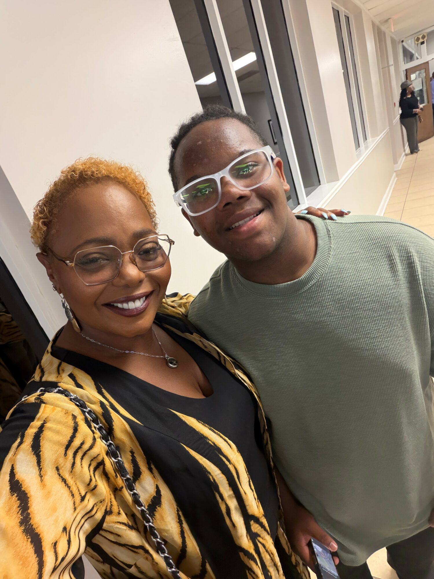 Two people smiling, one with short curly hair and glasses, the other with glasses and short hair, standing indoors.