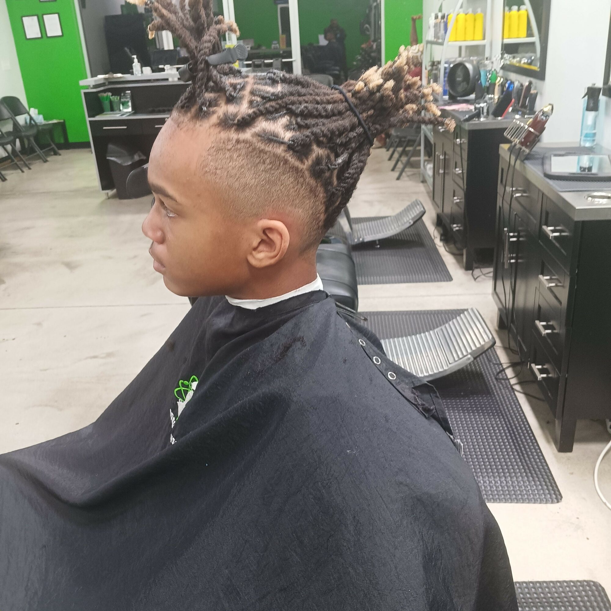 Person with braided hairstyle sitting in a salon chair, facing left, with salon equipment in the background.