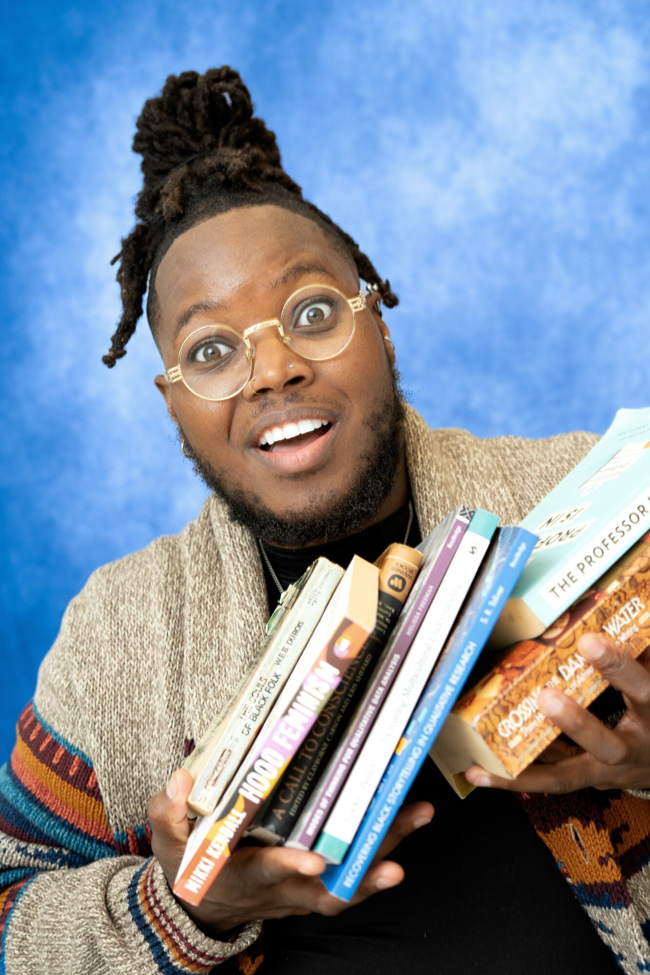 Person with glasses and dreadlocks holding a stack of books, smiling, against a blue background.