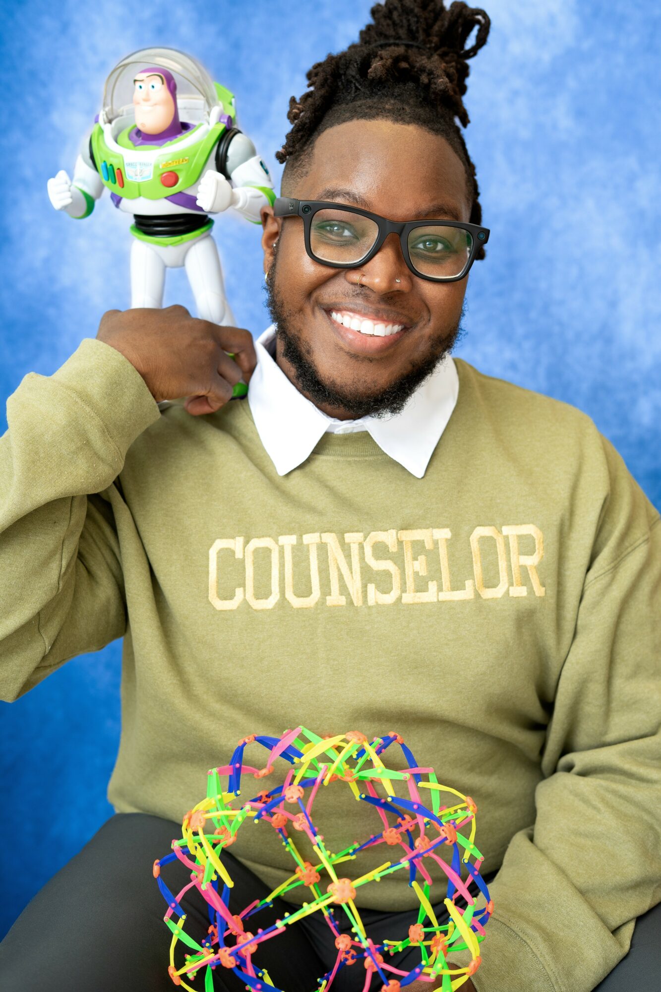 Man with glasses smiling, wearing a counselor sweater, holding a colorful toy, with a Buzz Lightyear figure on his shoulder, against a blue background.